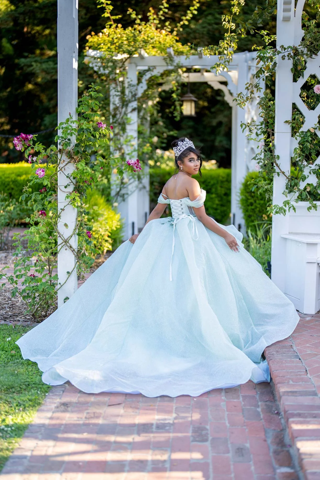 Young girl in a white ball gown and tiara sitting outdoors on a brick pathway surrounded by greenery and white garden structures.