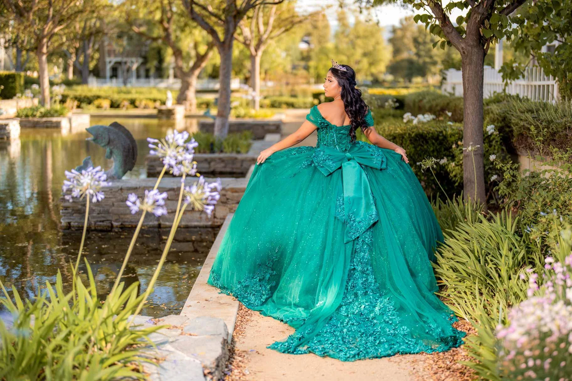 Young woman in an elaborate green ball gown with off-shoulder sleeves and a large bow, wearing a tiara, standing near a pond with a fish sculpture and purple flowers in a garden setting during daytime.