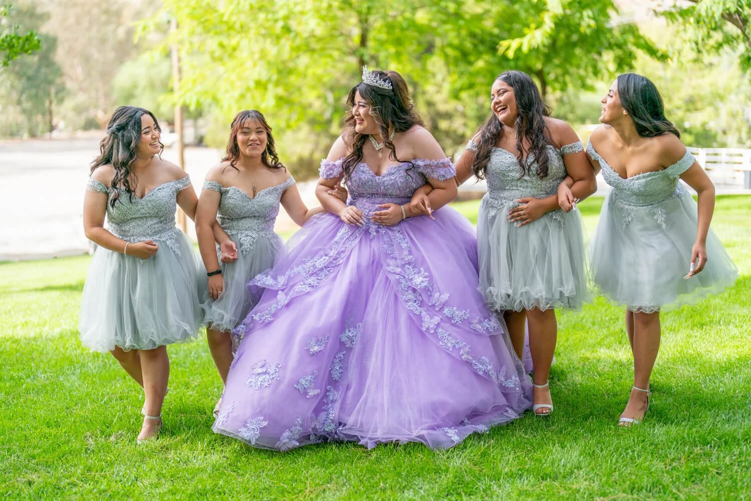A bride in a lavender dress with floral embroidery stands with five bridesmaids in silver-gray dresses outdoors on green grass, smiling and holding hands.