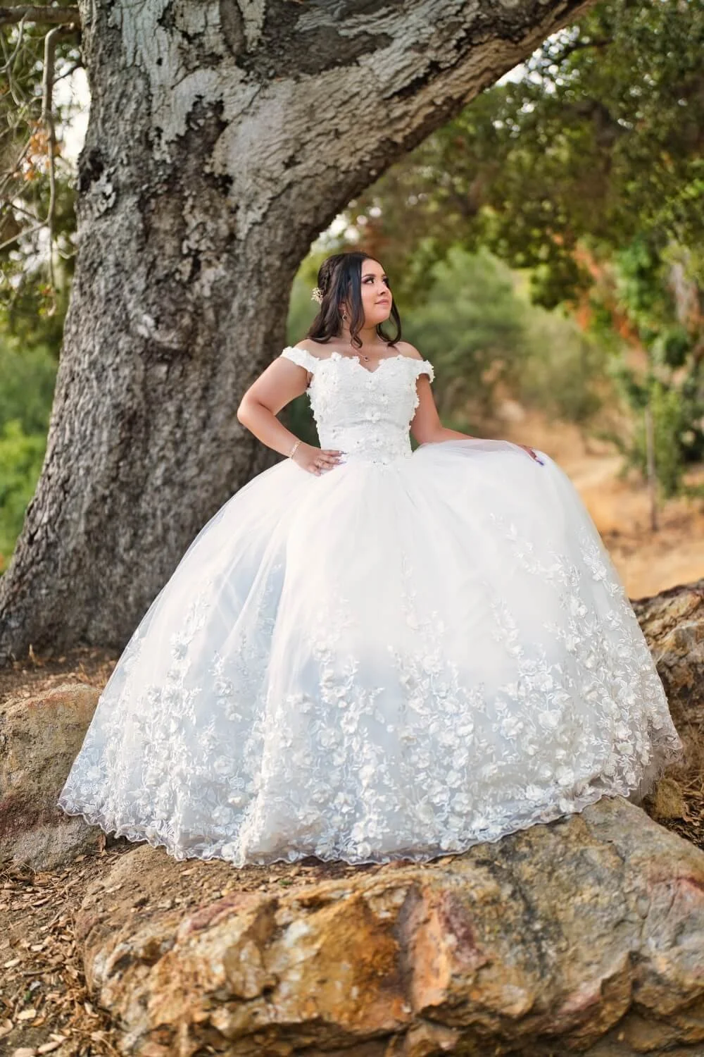 A woman in a white wedding dress standing outdoors in front of a large tree, with a scenic natural background of green foliage and a dirt path.