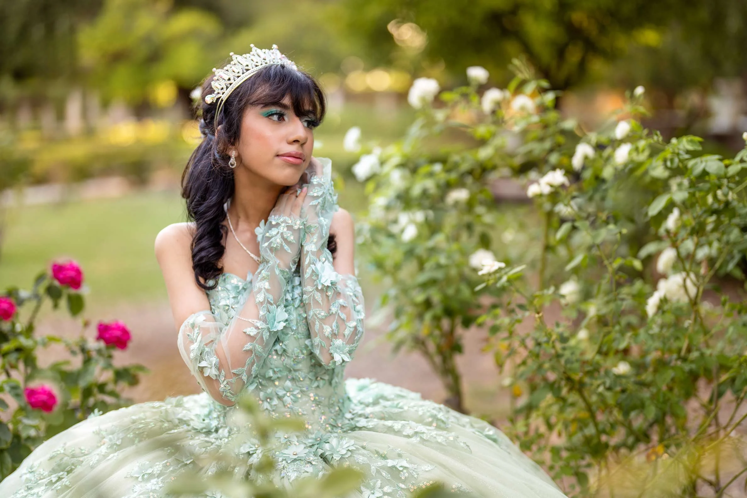 Young woman in a light green, floral embroidered gown with lace gloves, wearing a tiara and earrings, sitting among white and pink roses in a garden.