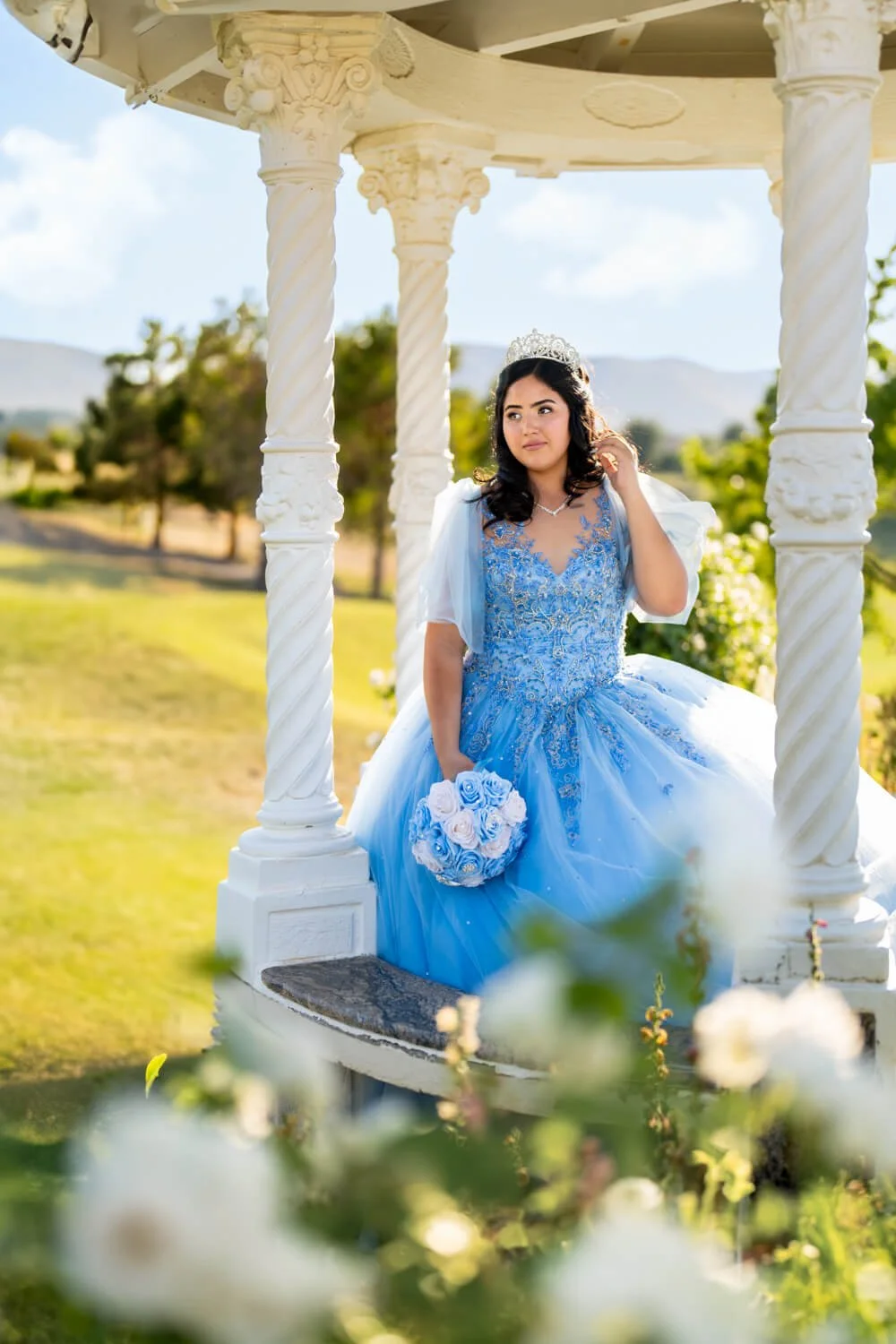 A young woman in a blue ball gown with intricate embroidery, wearing a tiara and holding a bouquet of white and blue roses, standing under a decorative white gazebo in a garden setting with trees and hills in the background.