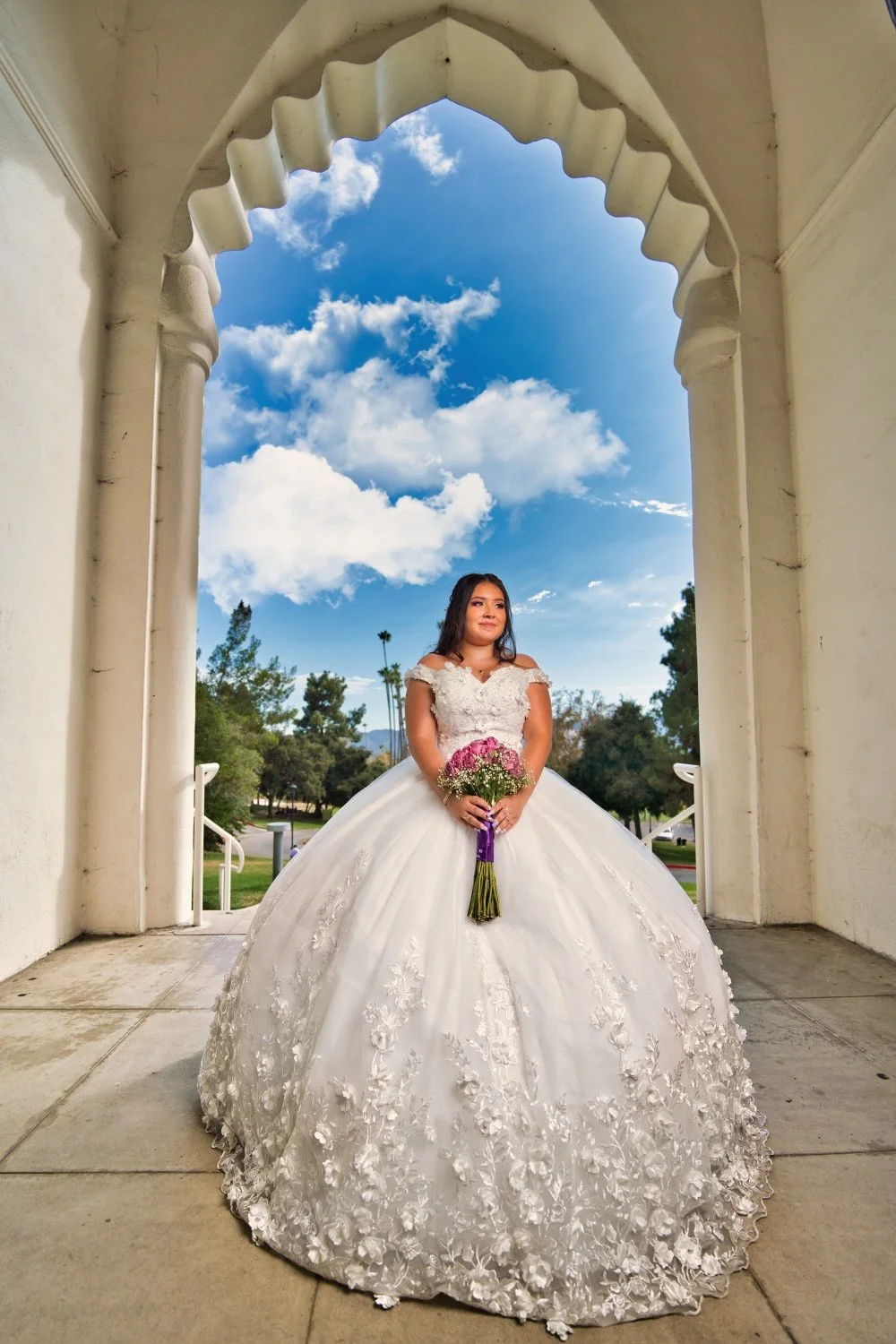 A bride in a white wedding gown holding a bouquet of pink and white flowers, standing under an archway with a blue sky and trees in the background.