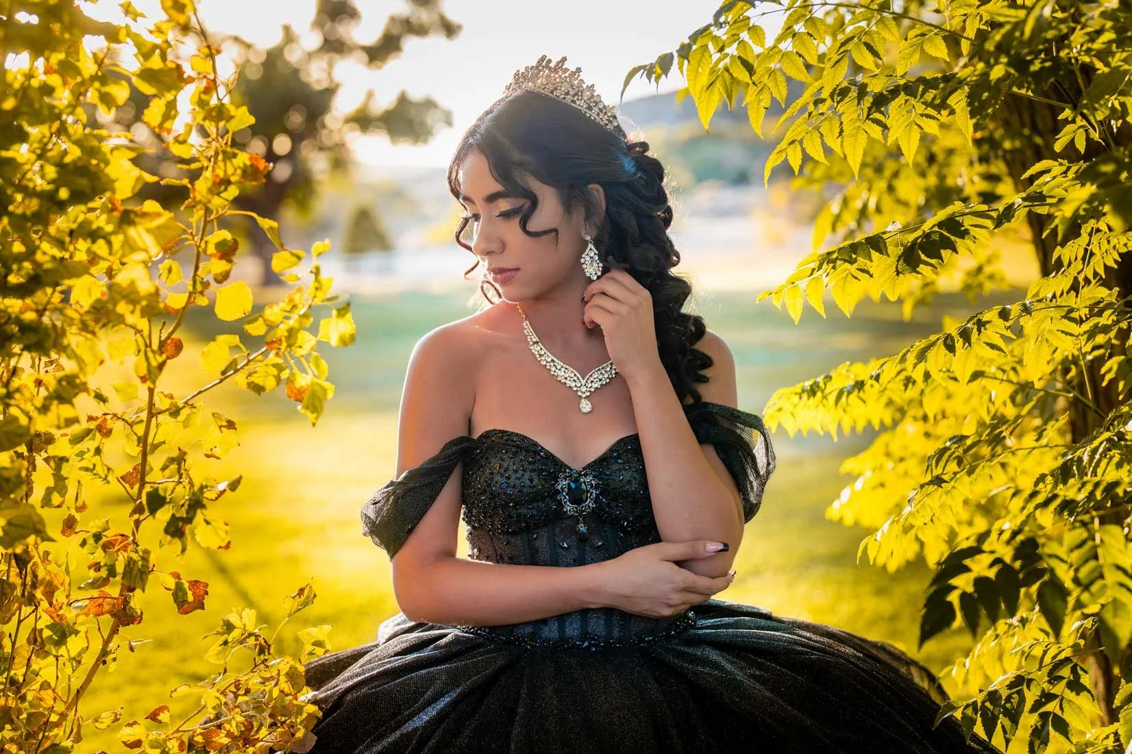 A young woman in a black, off-shoulder gown with jewelry, a tiara, and earrings, sitting outdoors among yellow-green leaves during golden hour.