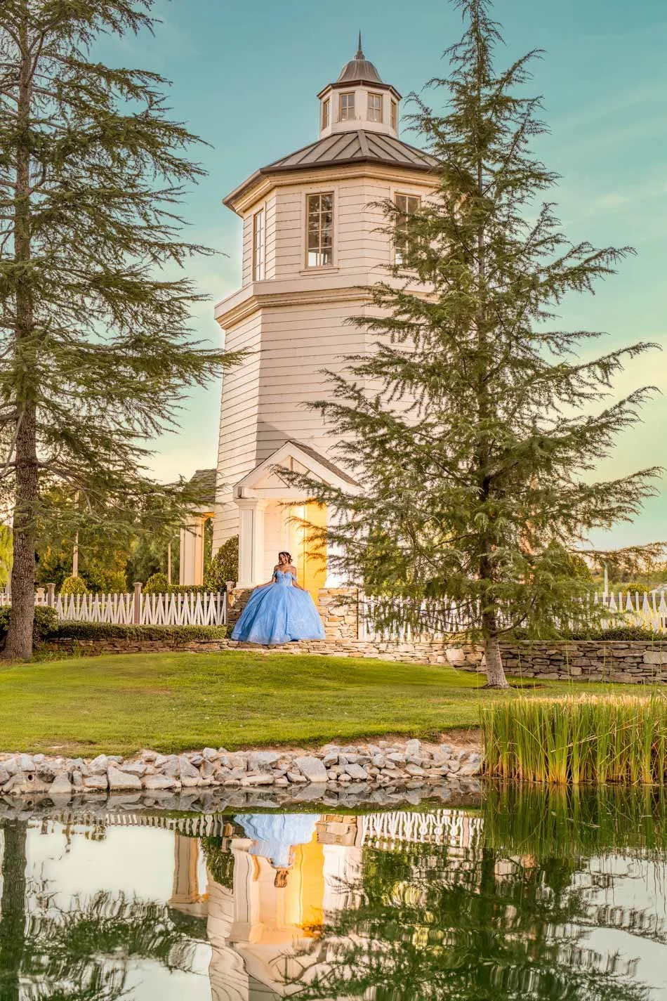 A woman in a blue ball gown standing outside a white wooden lighthouse near a pond with trees around.