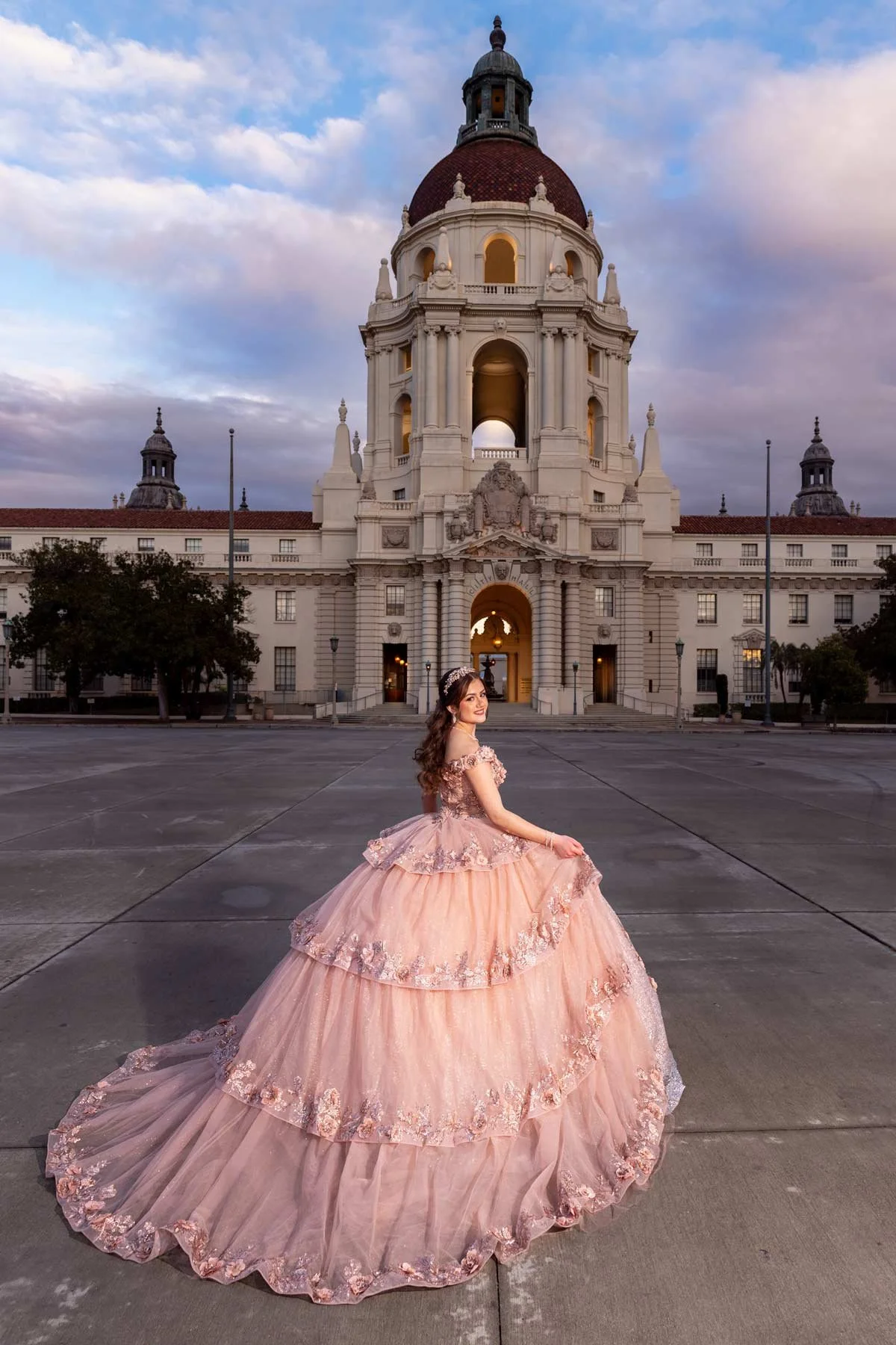 A woman in an elegant pink gown with floral embellishments standing in front of a large historic building with a domed roof during sunset.