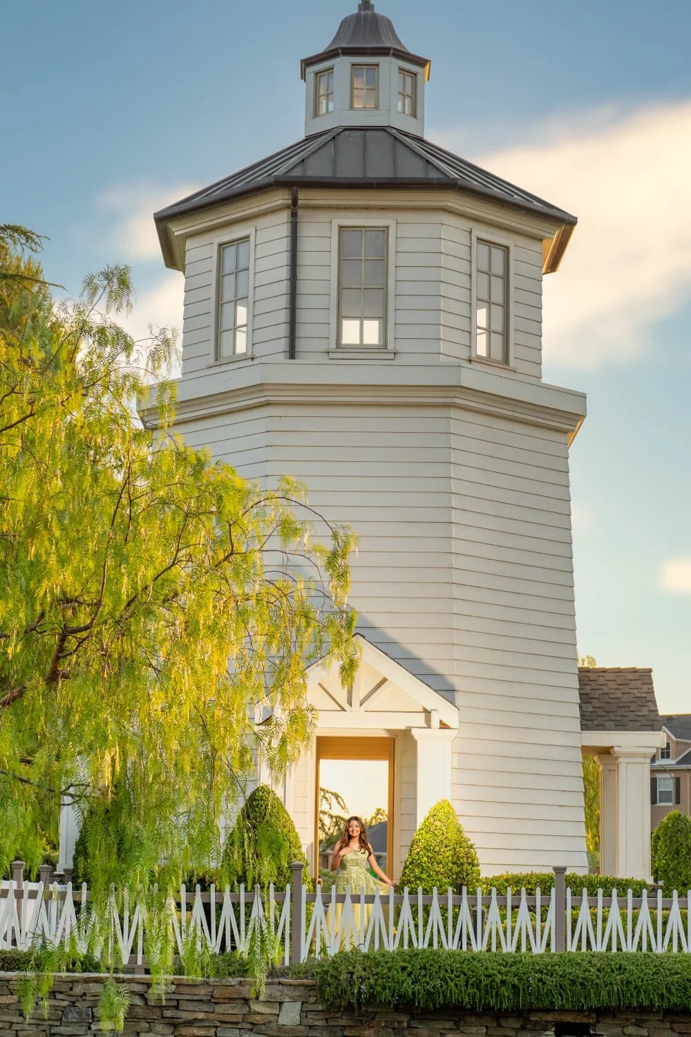 A woman in a yellow dress standing in front of a white wooden gazebo or tower with a pitched roof, surrounded by green trees and bushes, with a stone wall and white picket fence in the foreground, and a blue sky with some clouds in the background.