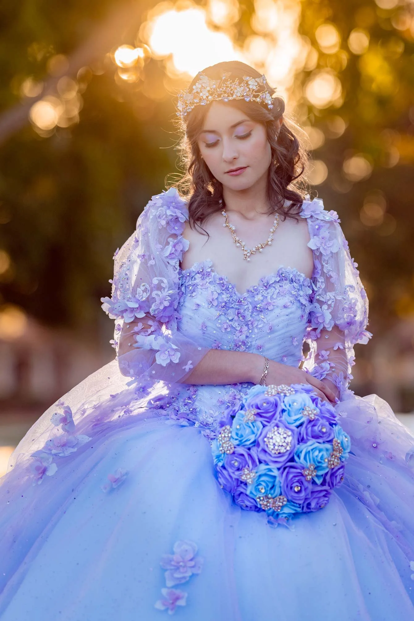 A woman in a lavender wedding gown with floral embellishments holding a bouquet of blue and purple roses. She is wearing a tiara and jewelry, with her eyes closed, outdoors at sunset.