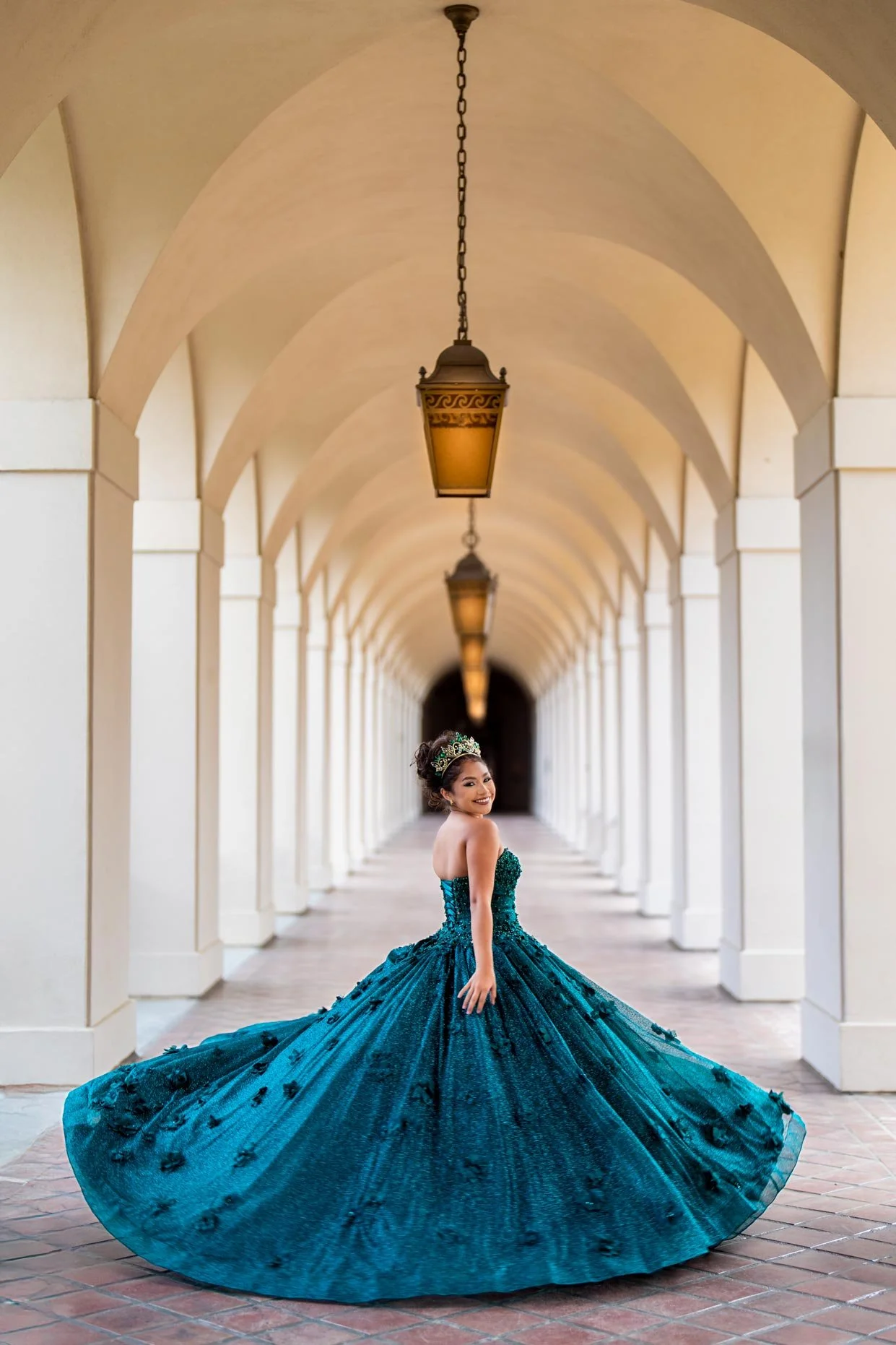 A woman in a teal ball gown with floral embellishments, wearing a sash and crown, poses in a covered walkway with arches and hanging lamps.