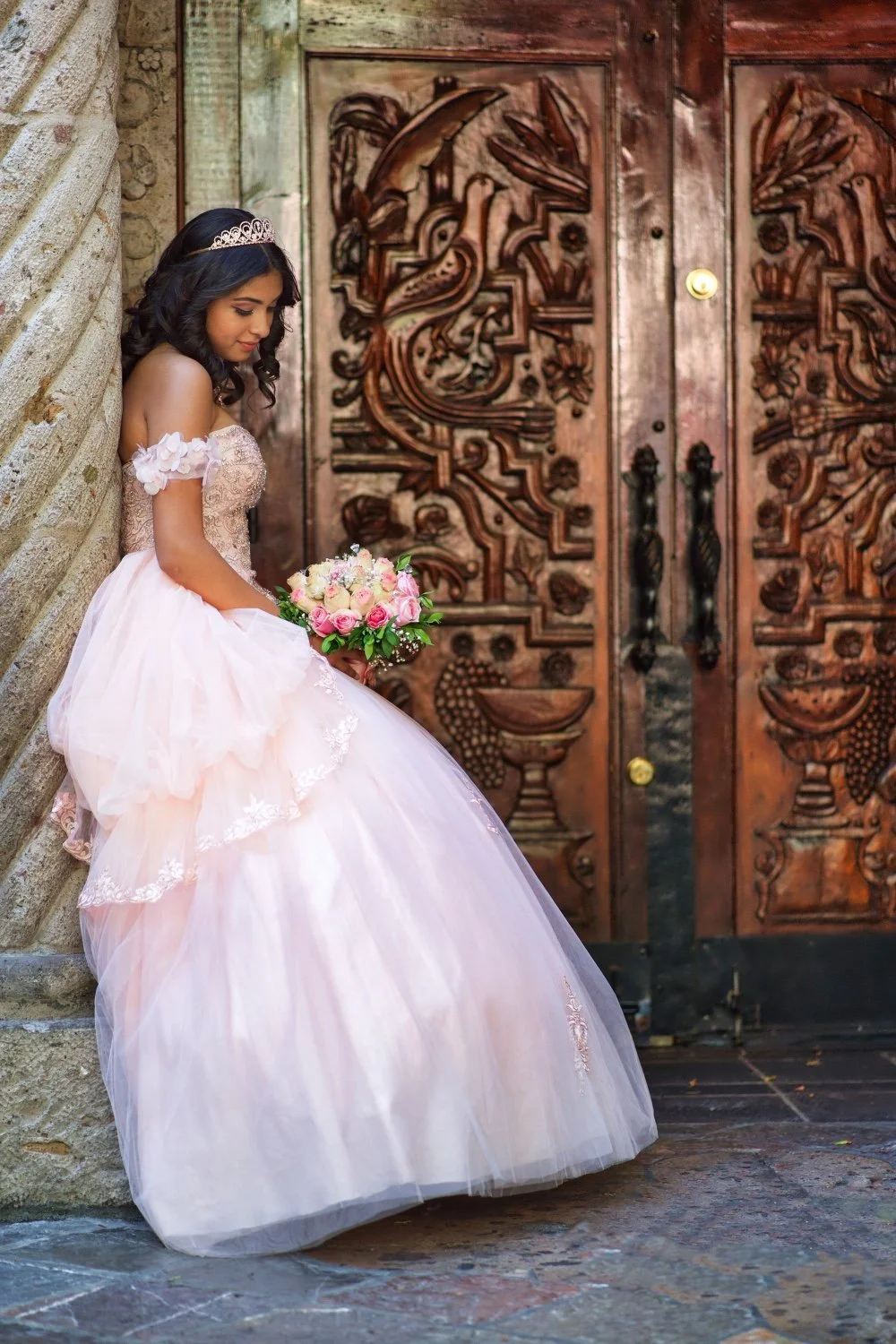 A young woman in a pink wedding dress leaning against a stone column, holding a bouquet of pink and white roses, with an intricately carved wooden door in the background.