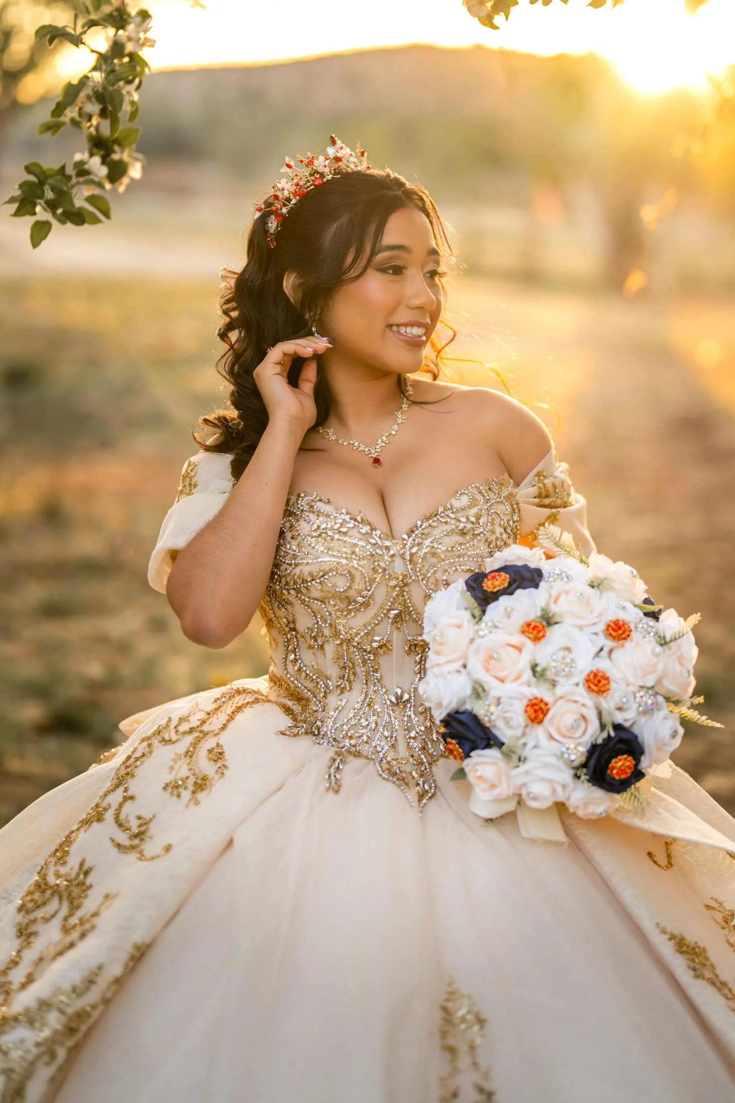 A woman in a wedding dress holding a bouquet of roses outdoors during sunset. She wears a tiara and necklace, smiling and touching her hair.