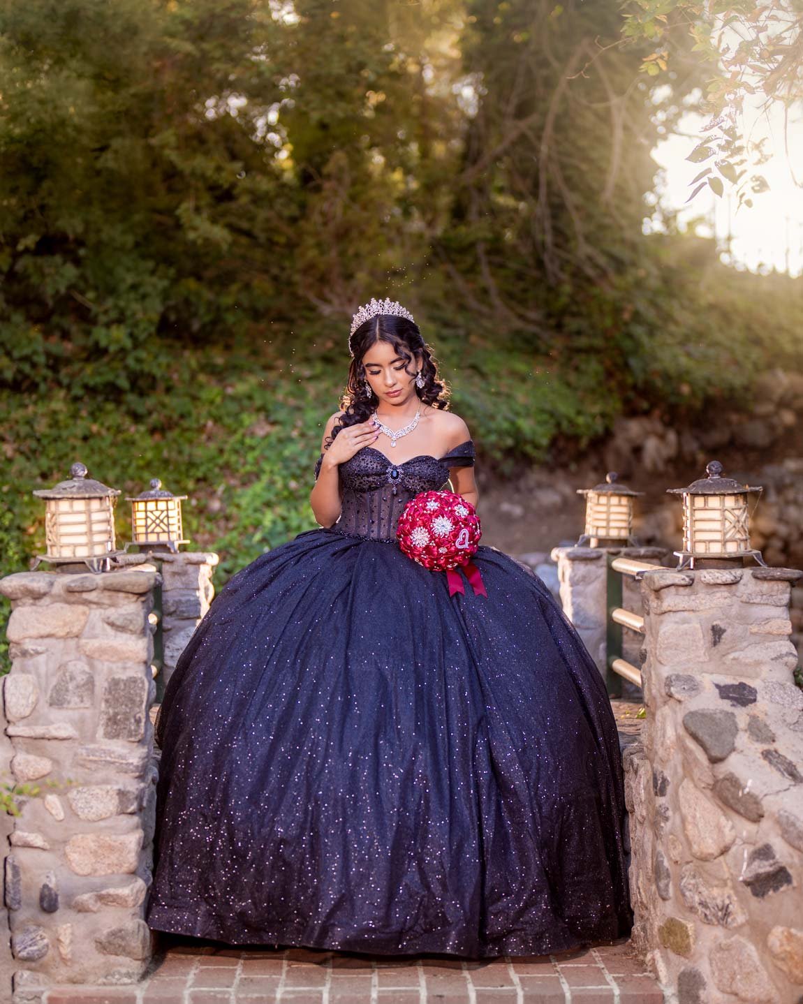 A woman in a black ball gown with glittering details, wearing a tiara and necklace, sitting on a stone bridge with lanterns, holding a bouquet of red flowers, outdoors with trees and sunlight in the background.