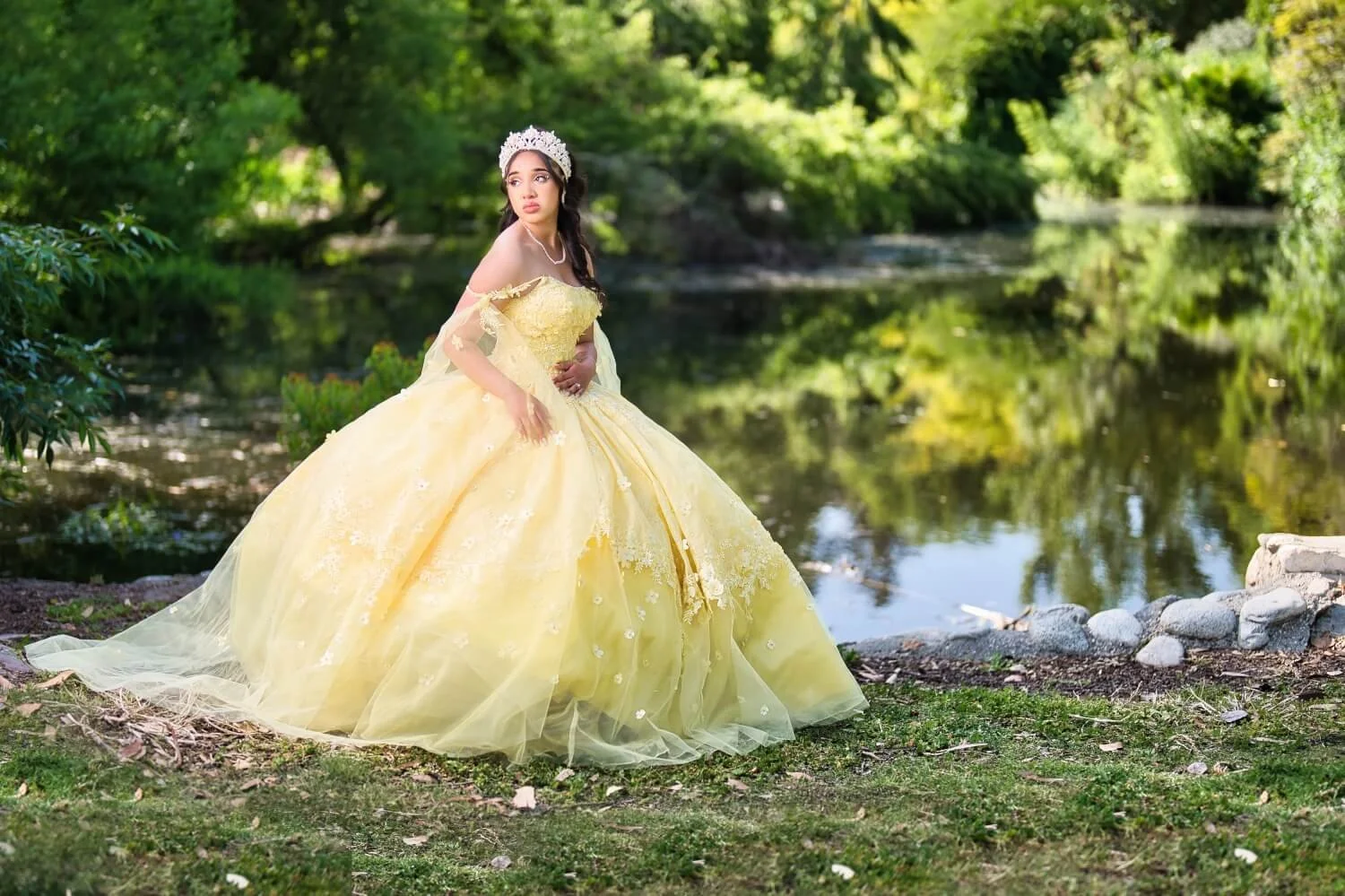 A young woman in a yellow ball gown with a tiara stands by a pond in a lush green park.
