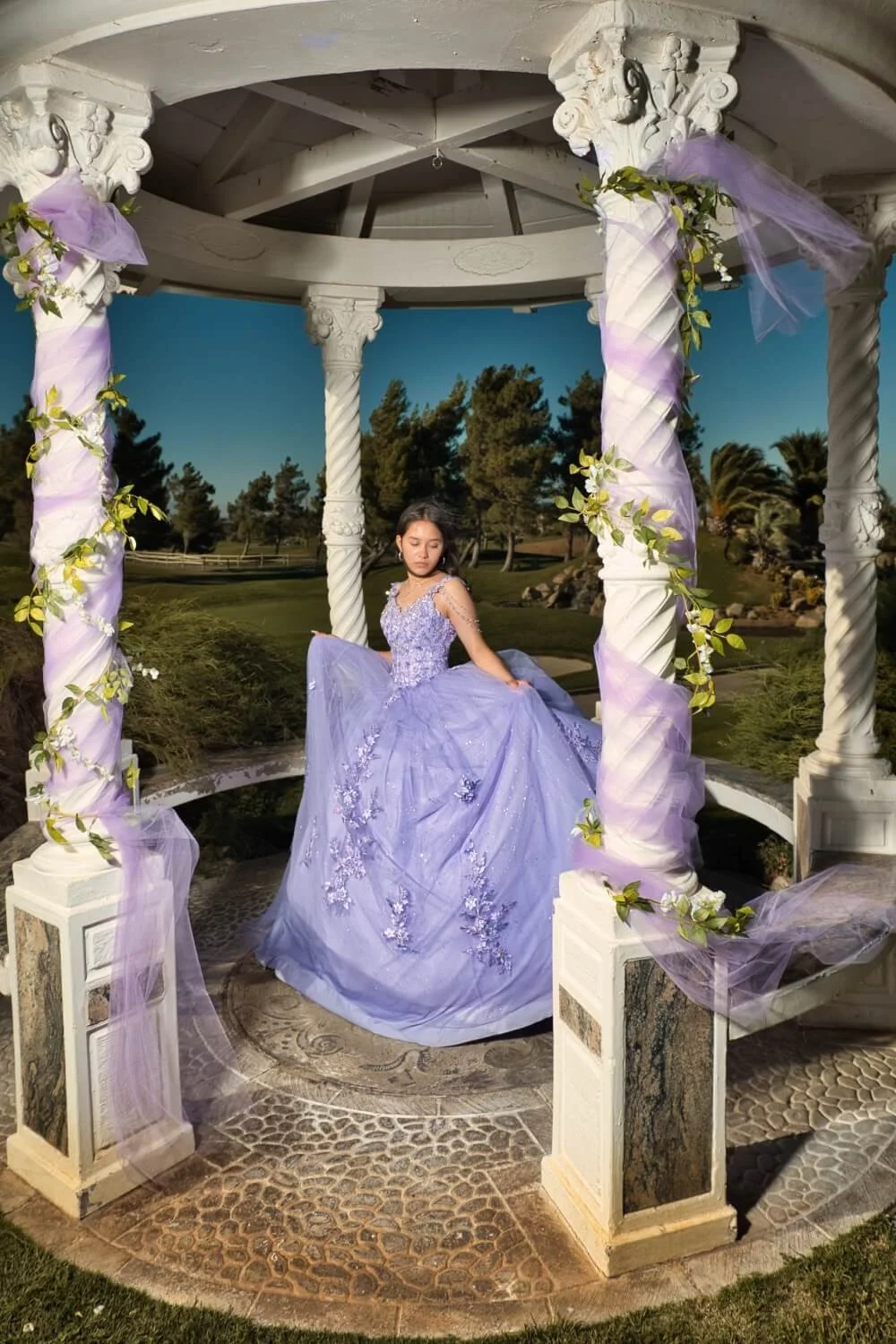 A young girl in a lavender ball gown sitting inside a decorated white gazebo in a garden setting.