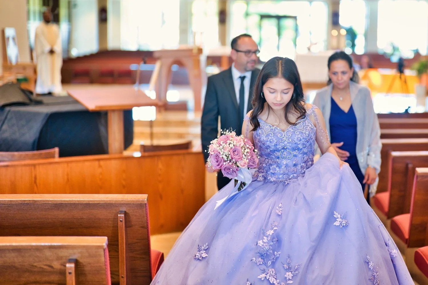 Young woman in a lavender gown holding a bouquet of pink and white roses walking down the aisle in a church with two adults following her.