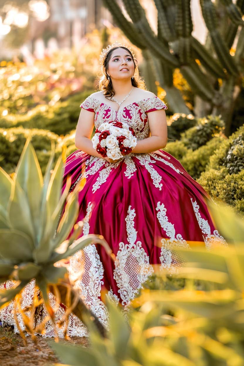 A woman in a burgundy and gold quinceañera dress holding a bouquet of roses, standing outdoors with cacti and desert plants.