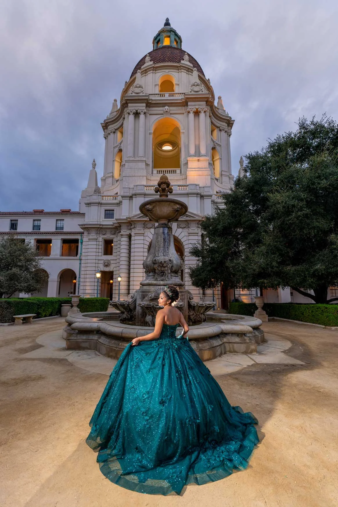 A woman in an elegant turquoise ball gown standing in front of a historic white building with a tower and domed roof during twilight.