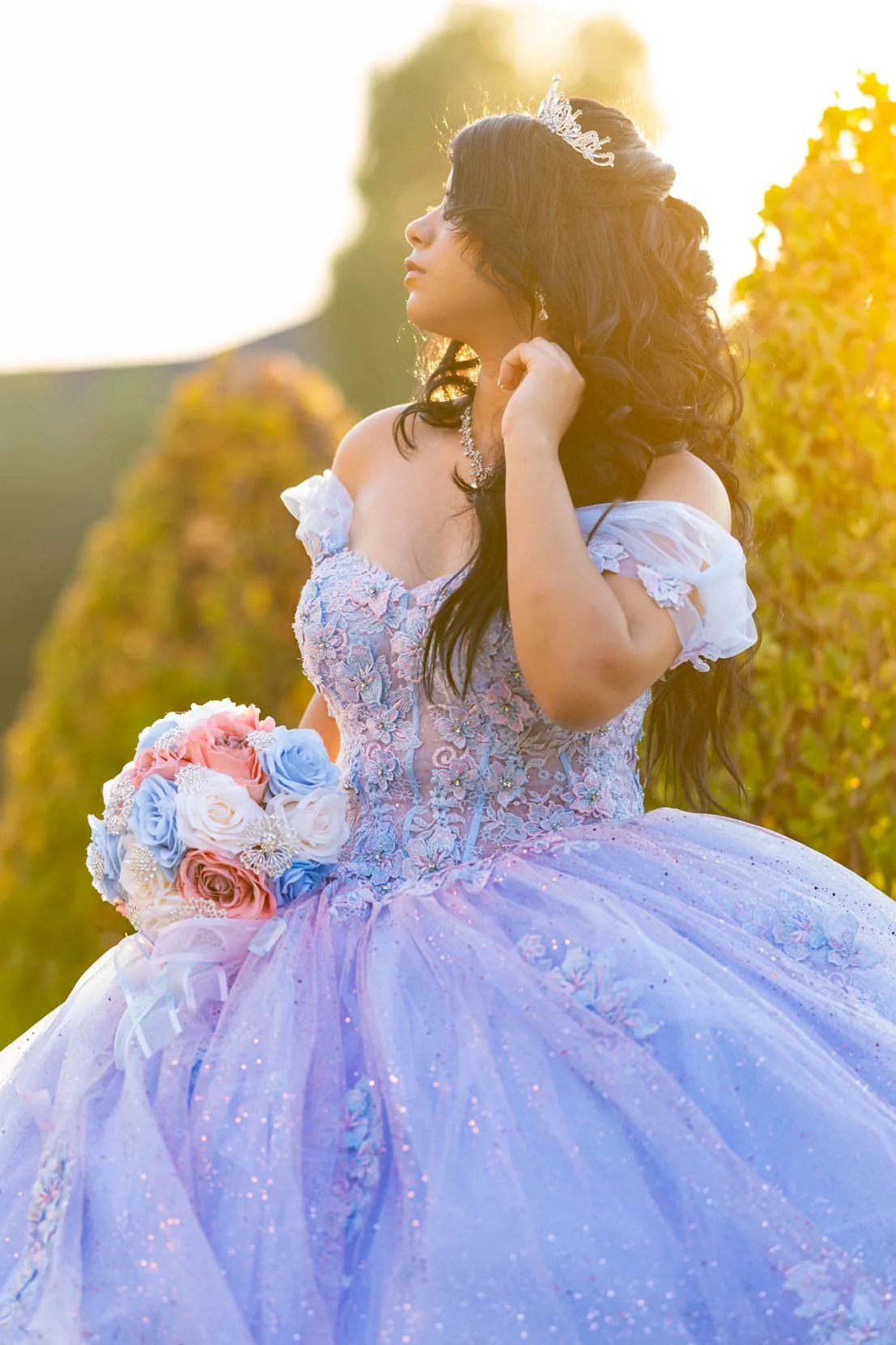 A woman in a light purple, off-the-shoulder ball gown with intricate embroidery and glitter detail, holding a bouquet of pink, white, and blue roses, and wearing a tiara and jewelry, standing outdoors during sunset.