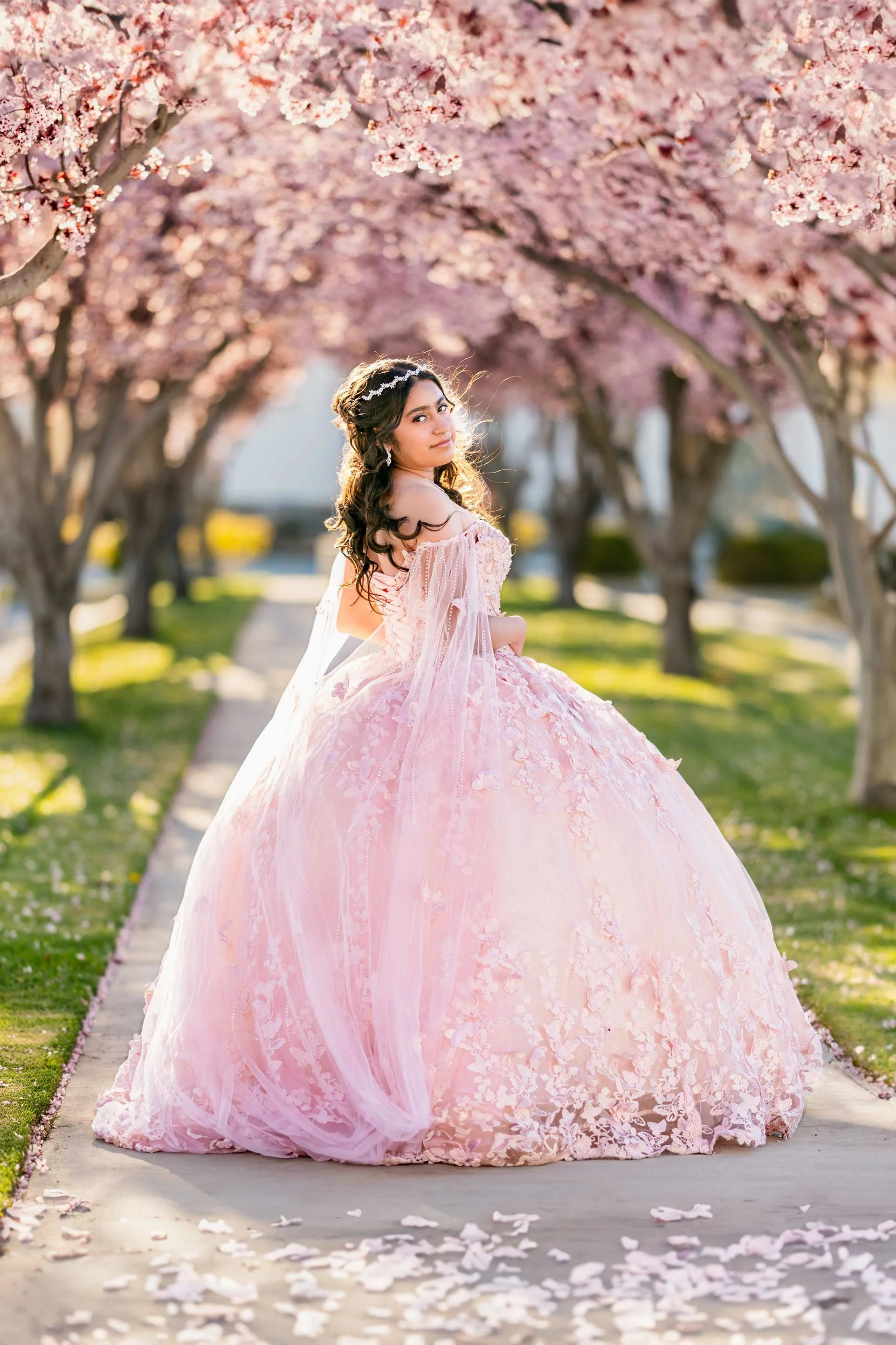 Young woman in a pink ball gown with floral embroidery sits on a walkway beneath blooming pink cherry blossom trees.