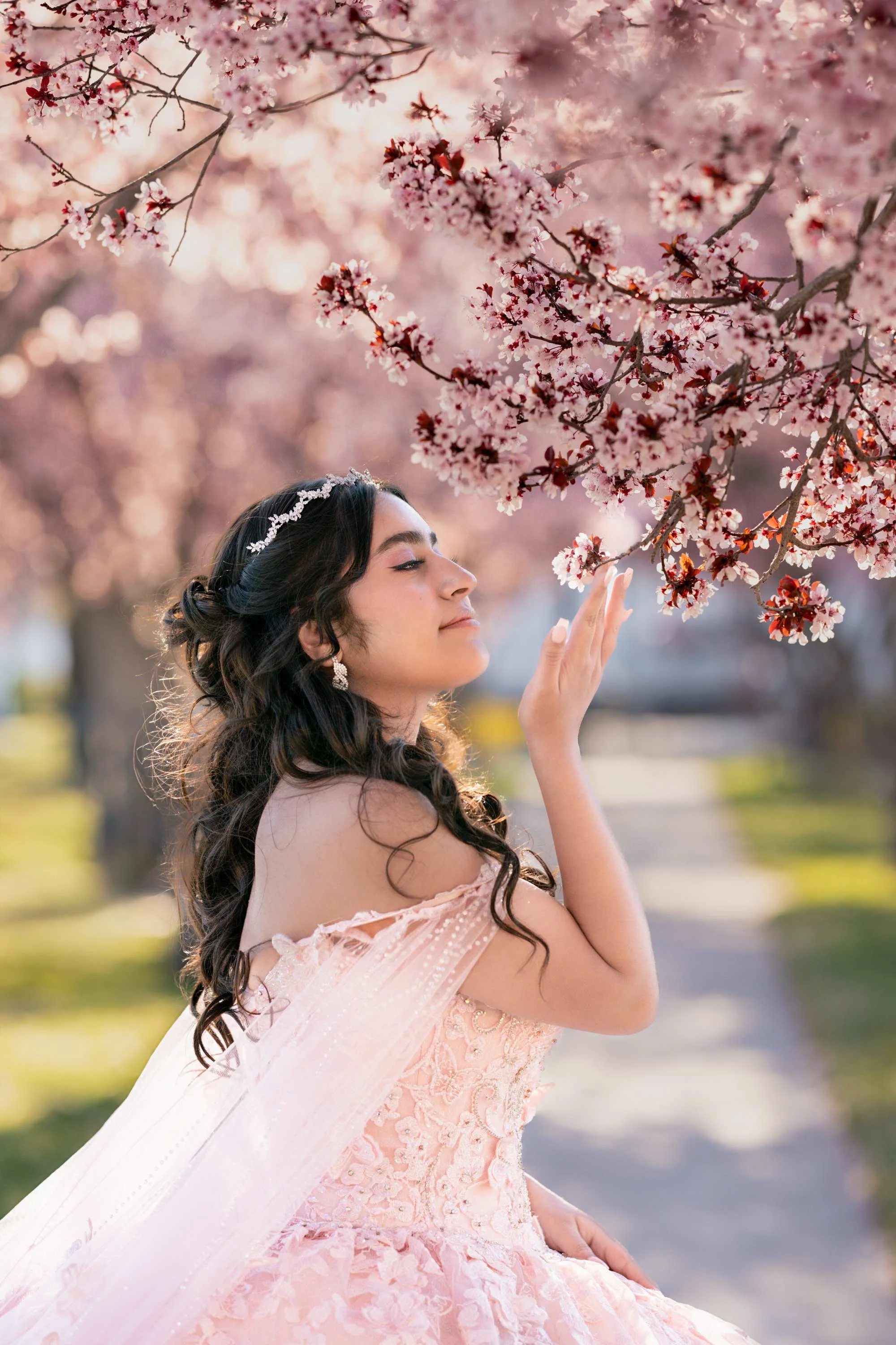 A young woman in a pink, off-shoulder dress with lace and embroidery, standing outdoors during spring, gently touching pink cherry blossoms on a tree branch with her eyes closed.