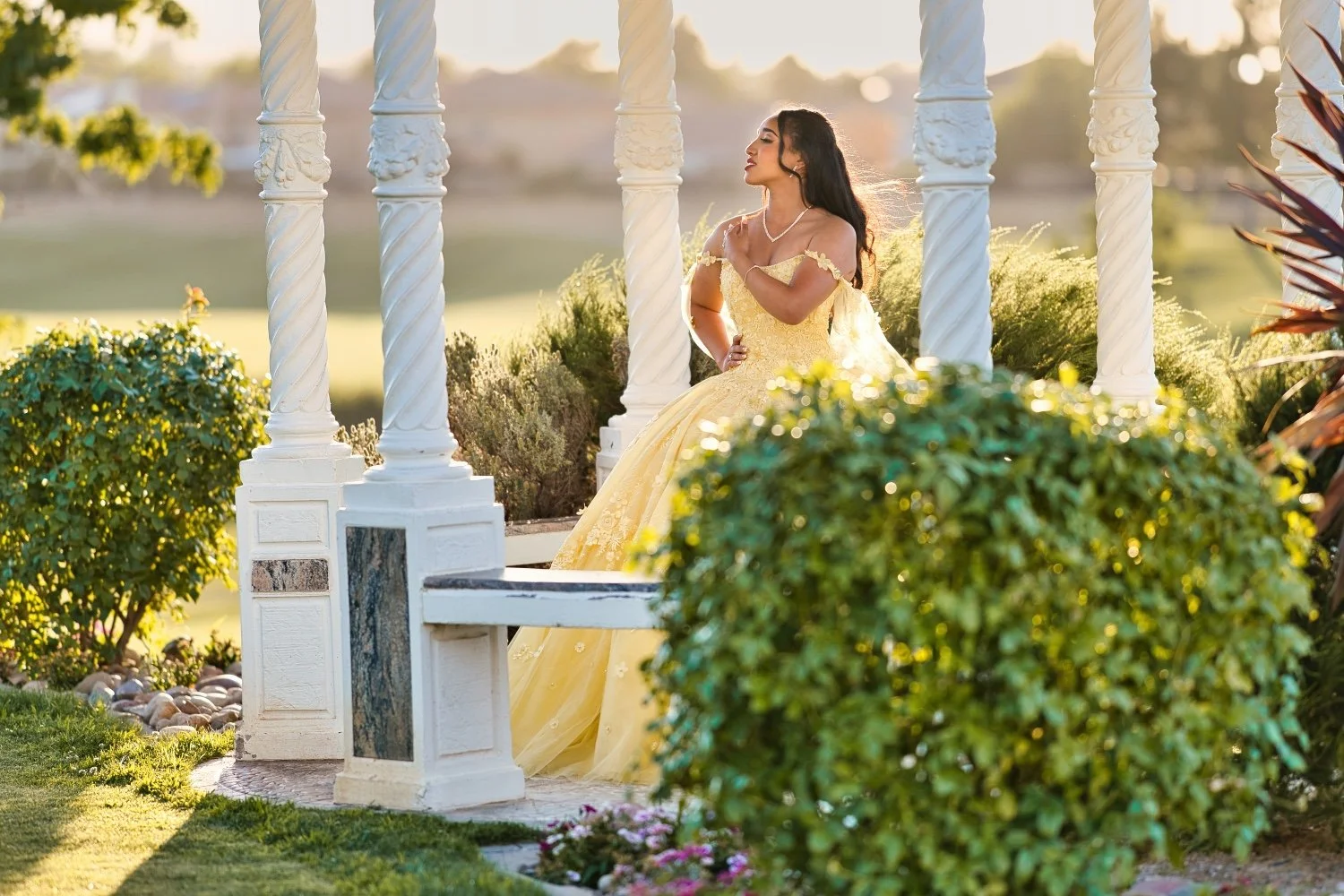 A woman in a yellow gown stands among white columns and greenery, looking thoughtfully to the side in an outdoor garden setting during sunset.