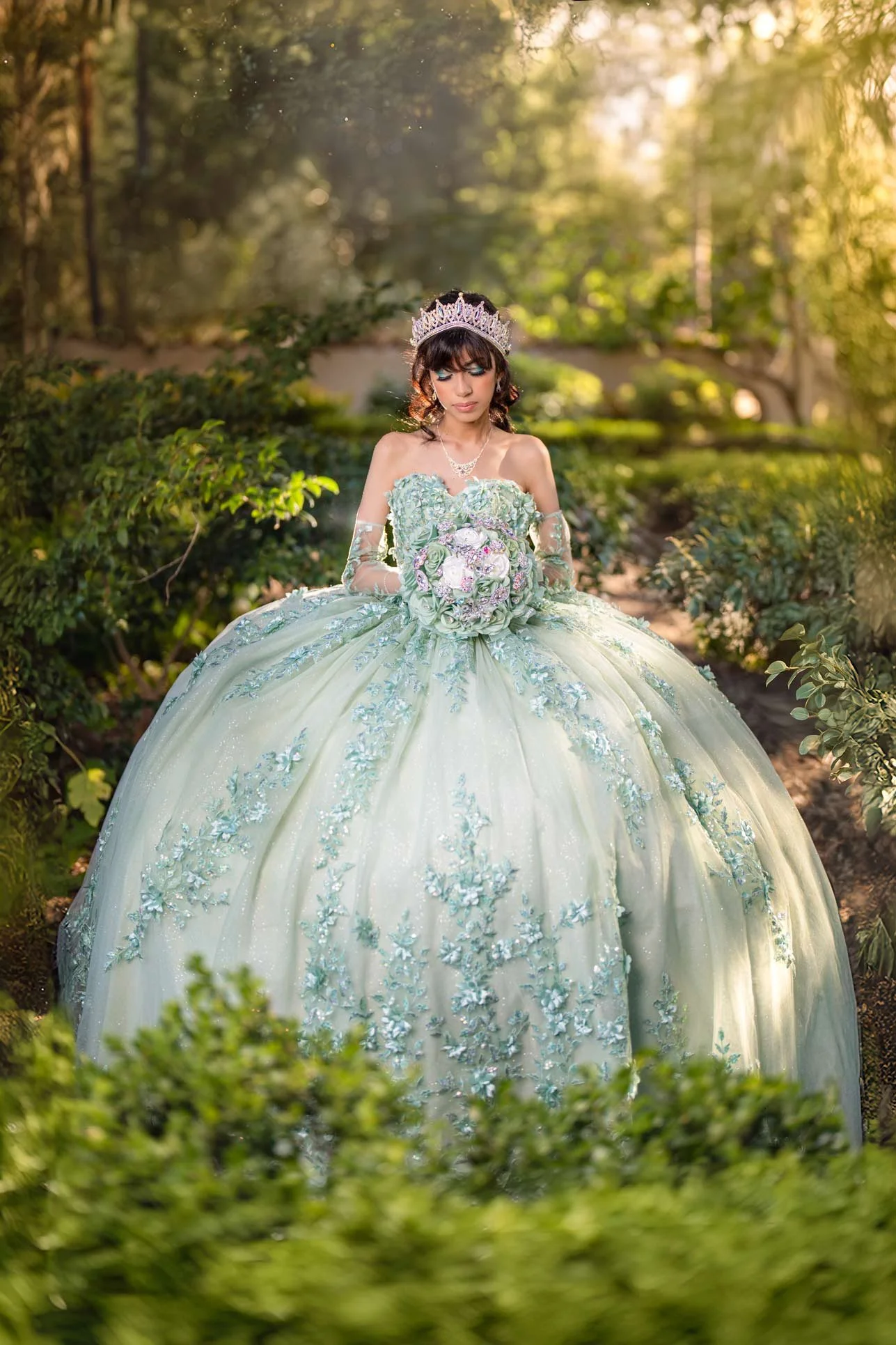 Young woman in an elegant, pastel green ball gown with floral embroidery, wearing a silver tiara and holding a bouquet of flowers, standing outdoors in a lush garden during golden hour.