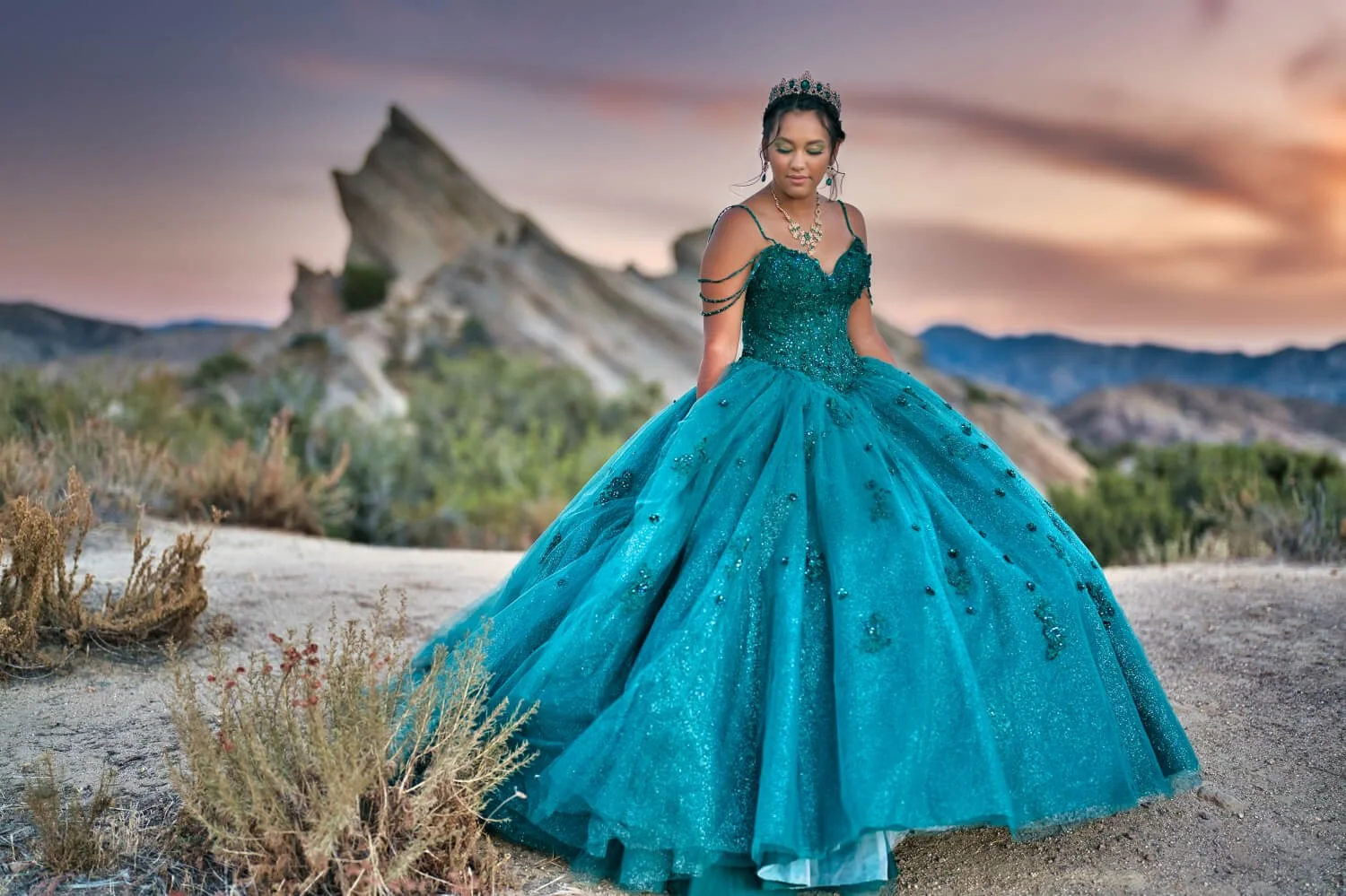 a young woman in an elaborate blue ball gown with a full skirt and off-the-shoulder design, standing outdoors in a desert landscape during sunset, wearing a tiara and jewelry
