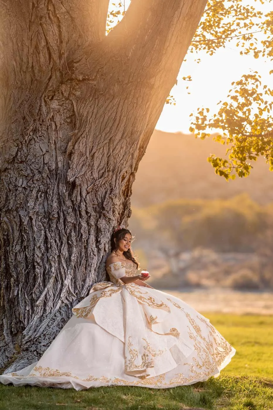 A woman in a white and gold elaborate gown leaning against a large tree trunk in an outdoor setting during sunset, holding a small cake.