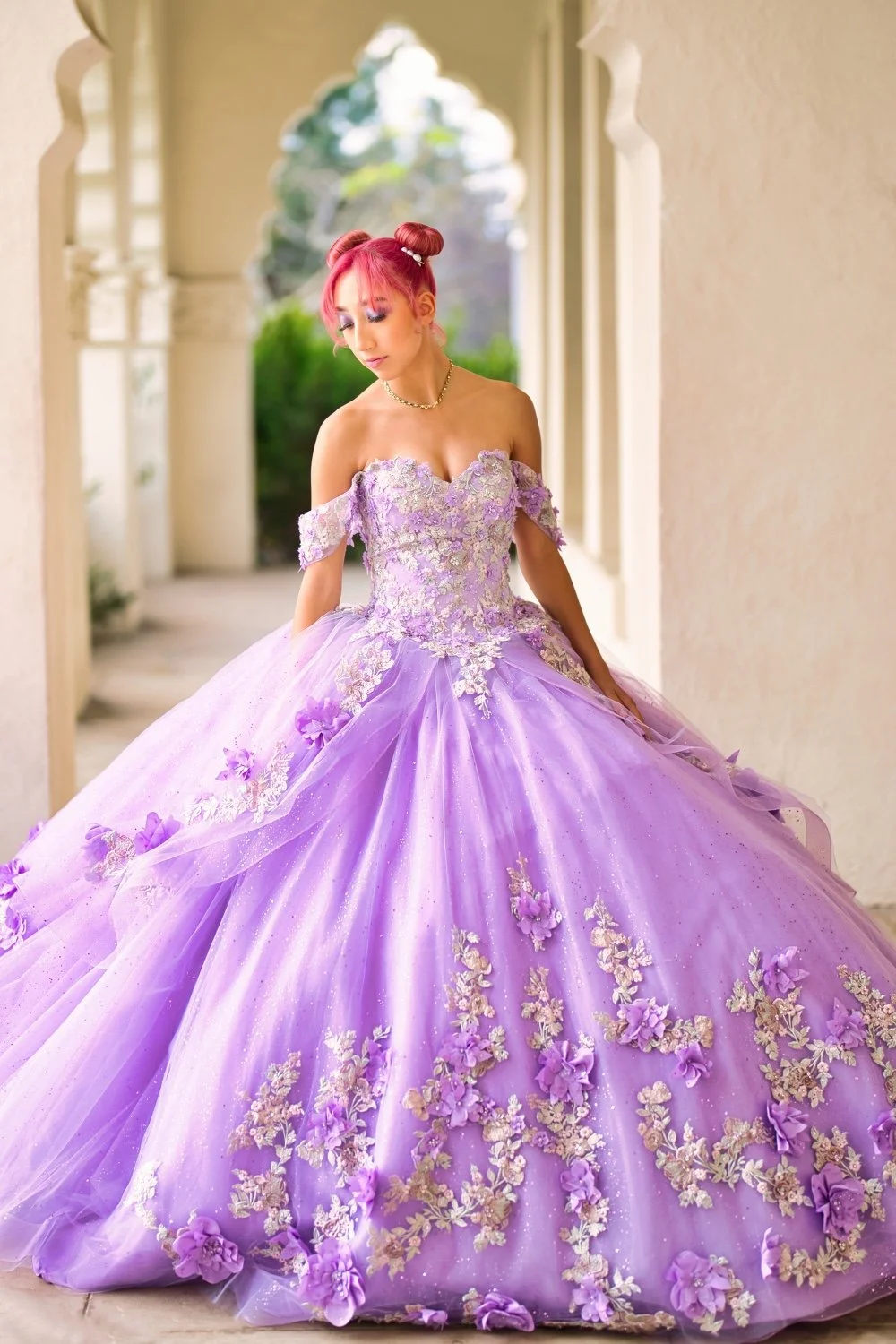 A young woman with pink hair styled in buns wearing a lavender off-the-shoulder quinceañera gown with floral embroidery and a full tulle skirt, standing in a corridor with arches.
