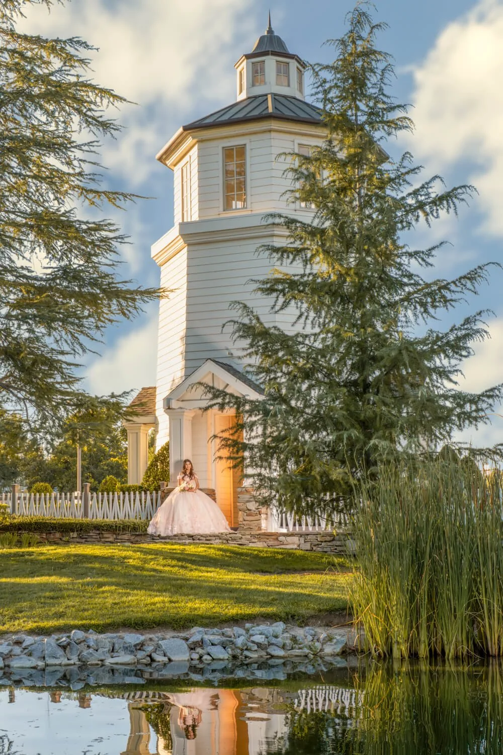 A woman in a white wedding dress standing on the steps of a white wooden lighthouse, surrounded by trees and a pond reflecting the scene.