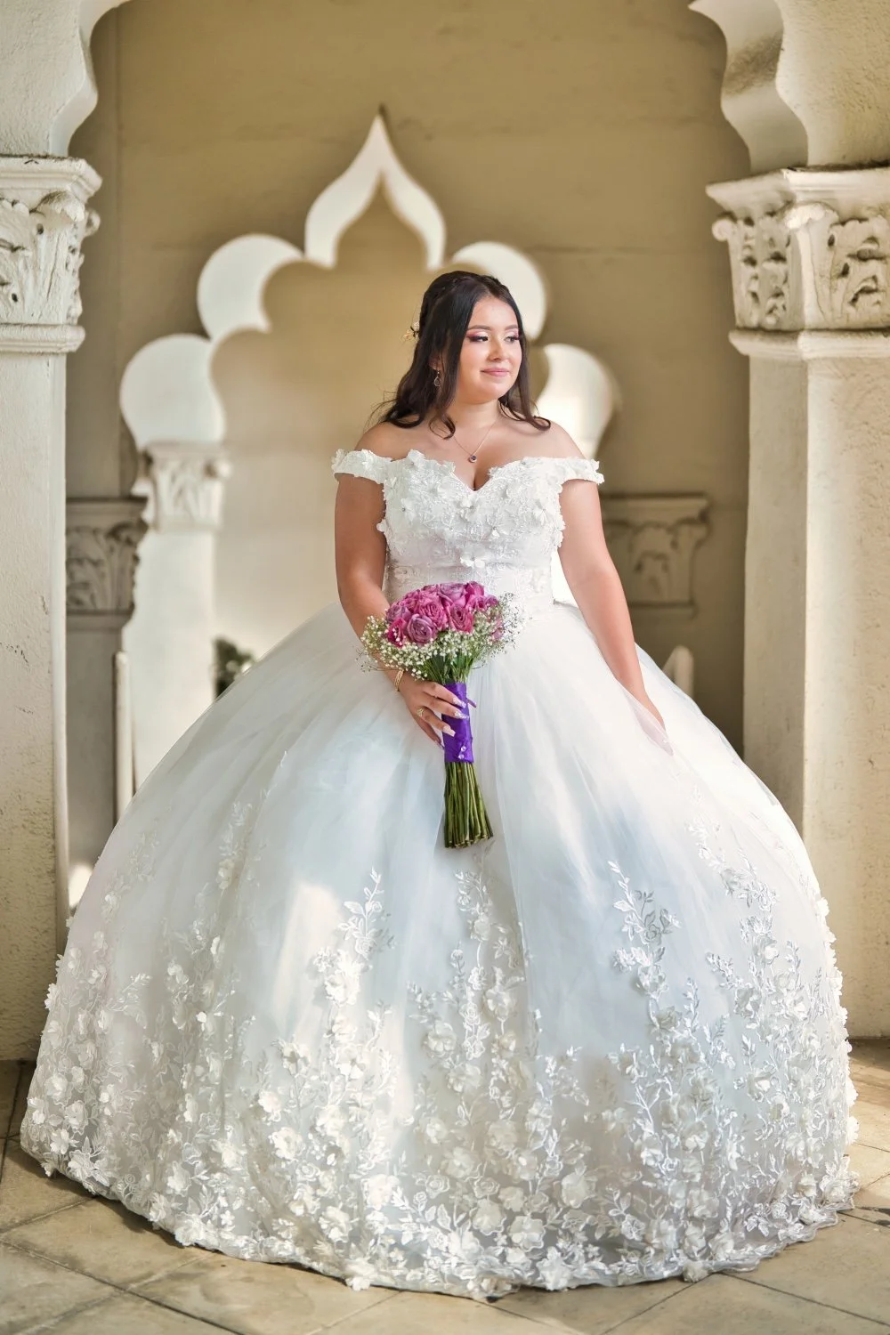 A bride in a white wedding gown with floral embroidery, holding a bouquet of pink roses, standing in front of a decorative archway.