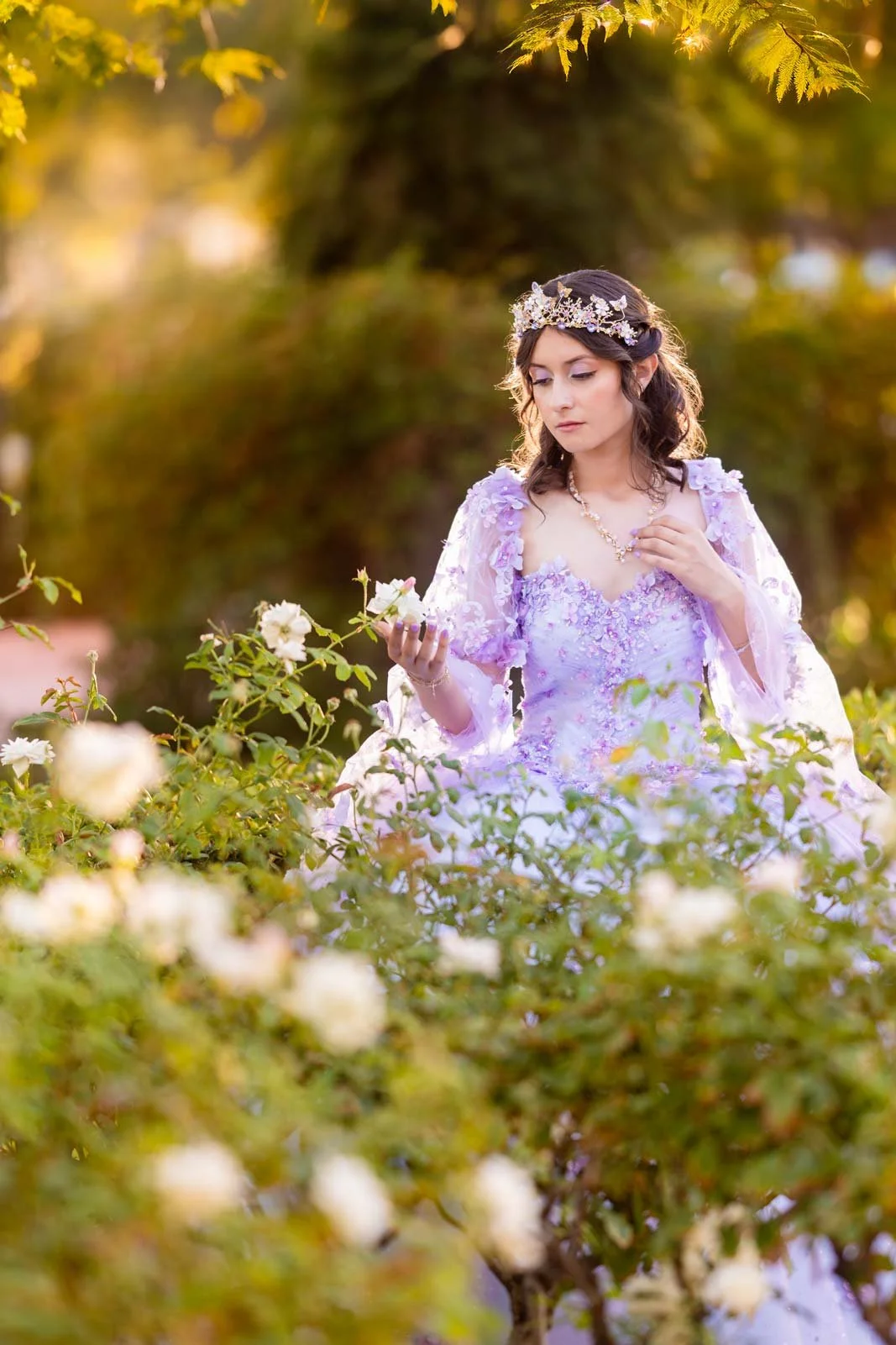 A young woman wearing a lavender dress with floral embroidery and a crystal tiara, is standing in a garden of white roses, holding a rose in her right hand, with sunlight filtering through green leaves in the background.