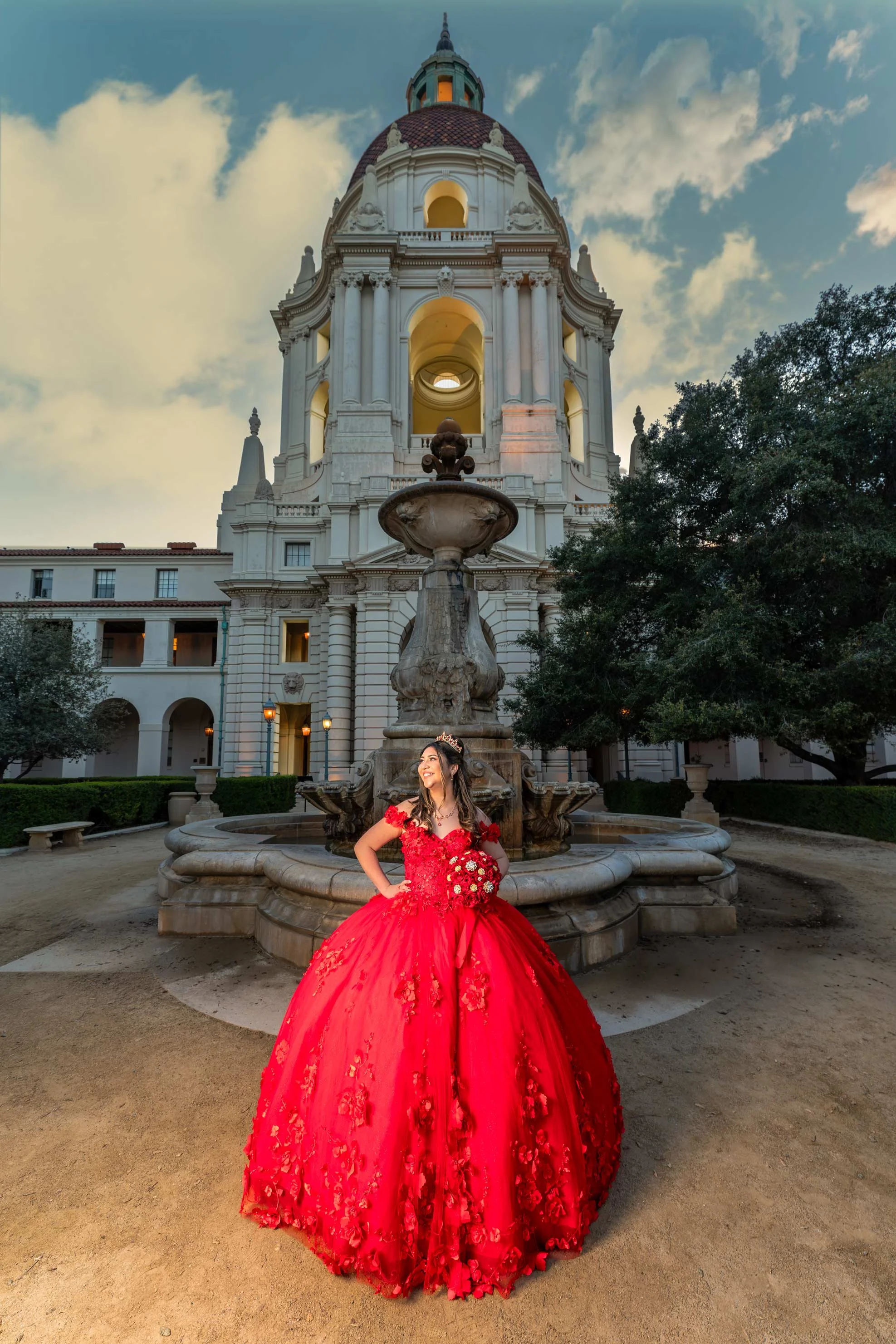 A woman in a bright red, detailed gown with floral embellishments, standing in front of a large fountain and historic building with a tower and domed roof at dusk, holding a bouquet and smiling.