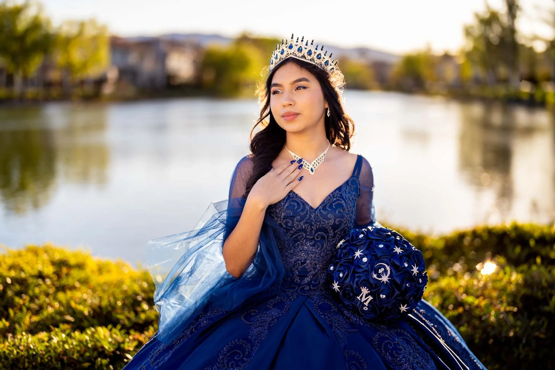 Young woman in a blue dress with a tiara and matching jewelry, holding a bouquet, outdoors near a lake with trees and buildings in the background.