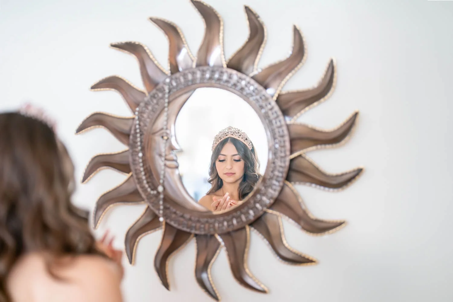 A woman with long wavy hair and makeup wearing a tiara looks at her reflection in a decorative sun-shaped mirror with a face in the center.