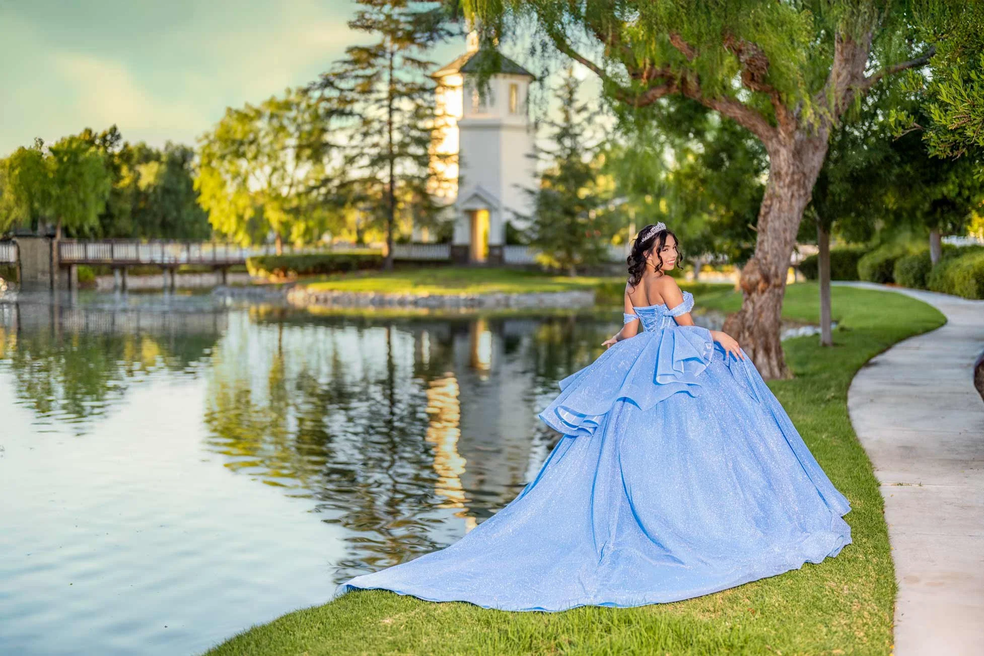 A woman in a sparkling blue gown and tiara sitting by a lake with a white building and trees in the background.