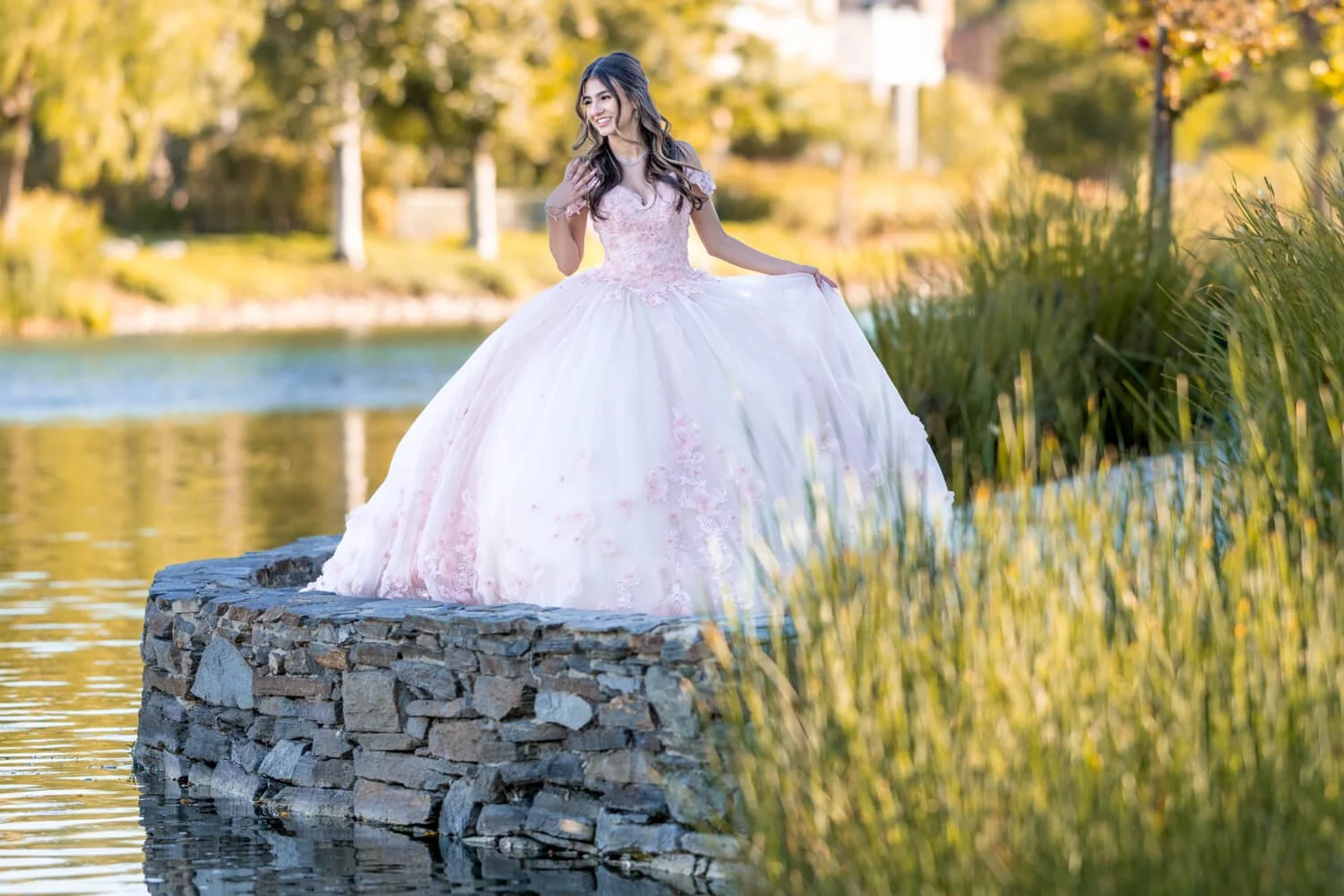 A young woman in a pink and white ball gown standing on a stone platform by a lake, surrounded by autumn-colored trees.
