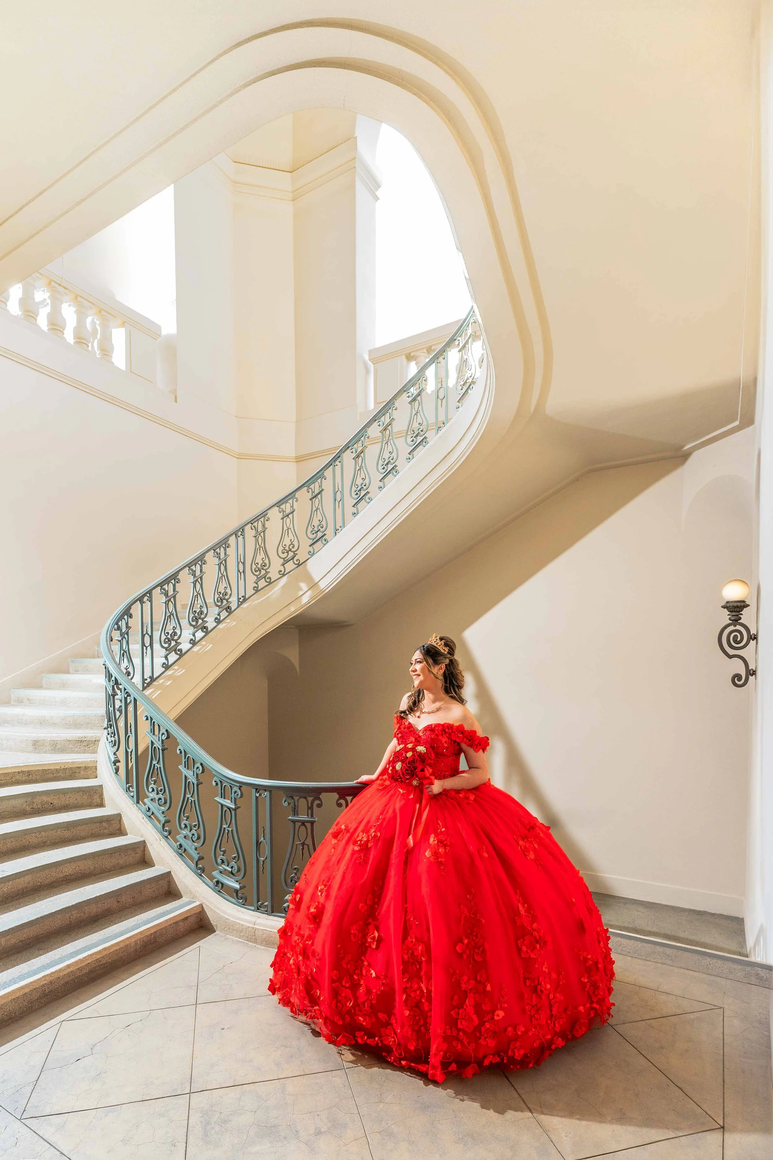 A woman in a bright red, off-the-shoulder, ball gown style wedding dress with floral embellishments, standing at the bottom of a curved staircase in a grand interior space, holding a bouquet of red roses and wearing a tiara.