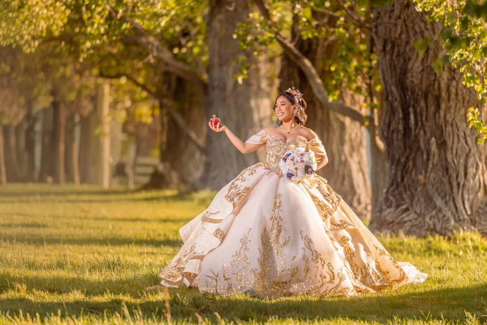A woman in an ornate golden and cream wedding gown holding a bouquet of flowers in a park with large trees and green grass.