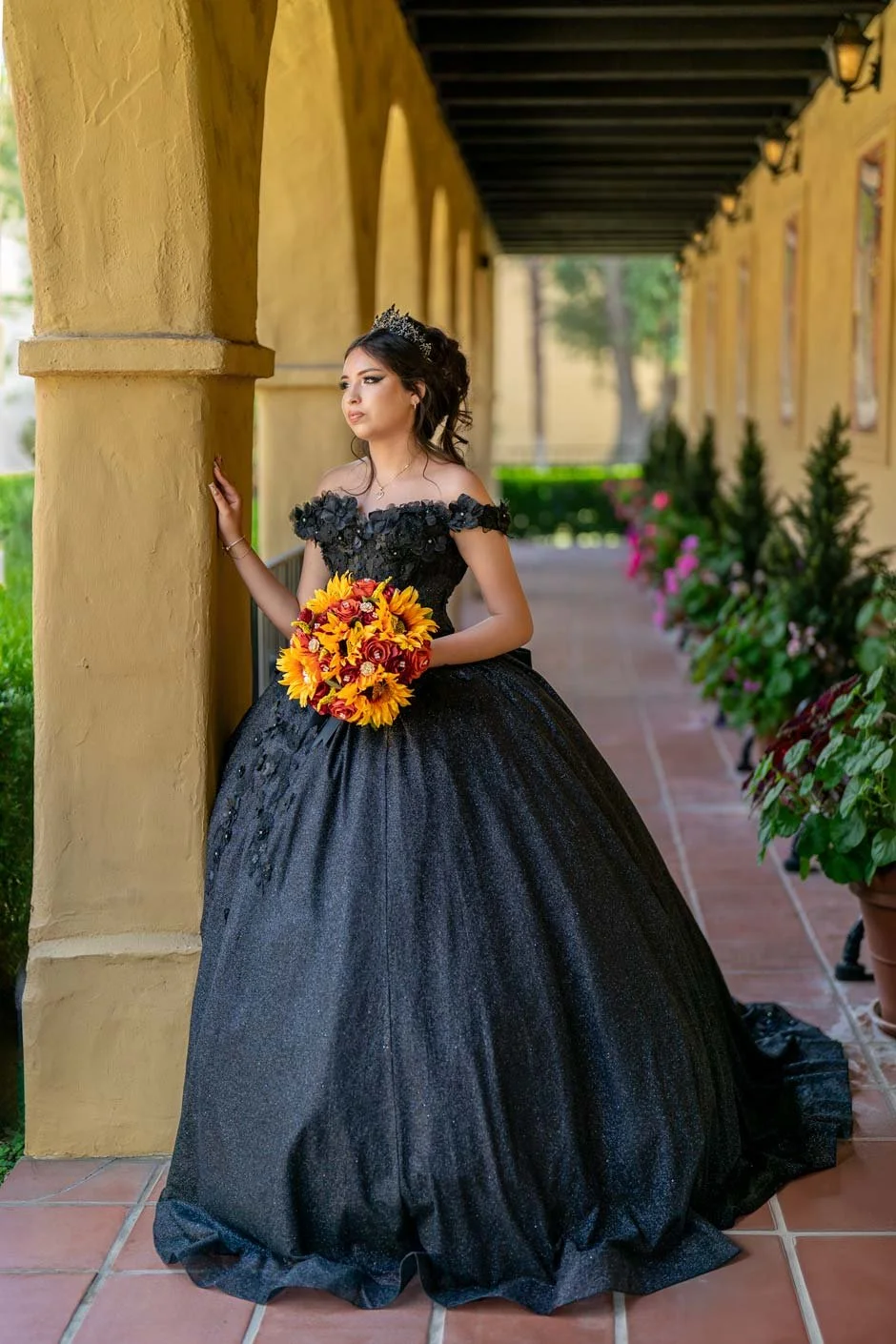 Young woman in a black ball gown holding a bouquet of sunflowers and red roses, standing in a corridor with yellow arches and potted plants.