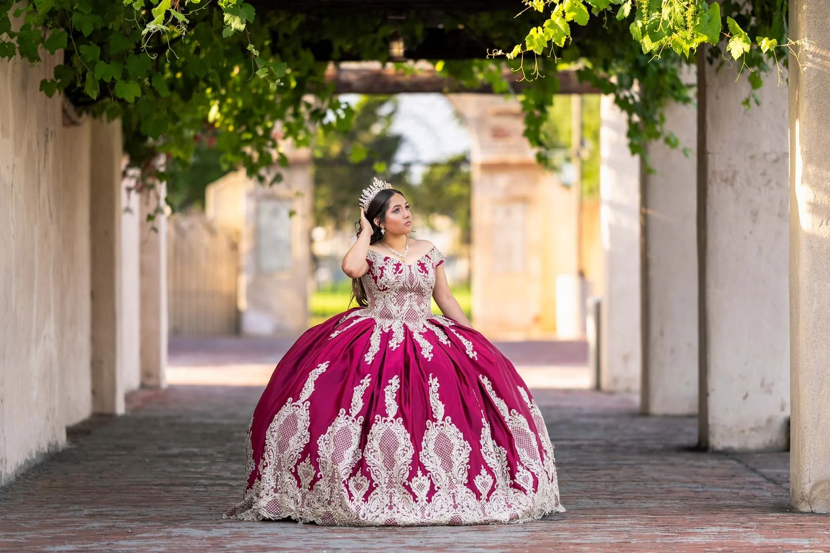 Young woman in a pink and gold ball gown with intricate embroidery, wearing a tiara and earrings, standing under a tree archway with greenery, on a brick pathway in an outdoor setting.