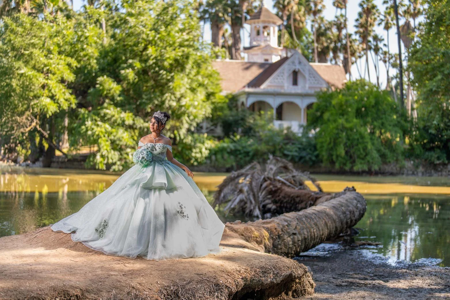 A woman in an elegant wedding dress holding a bouquet, standing on a sandy area by a river with a large fallen tree, greenery, and a house with a tower in the background.