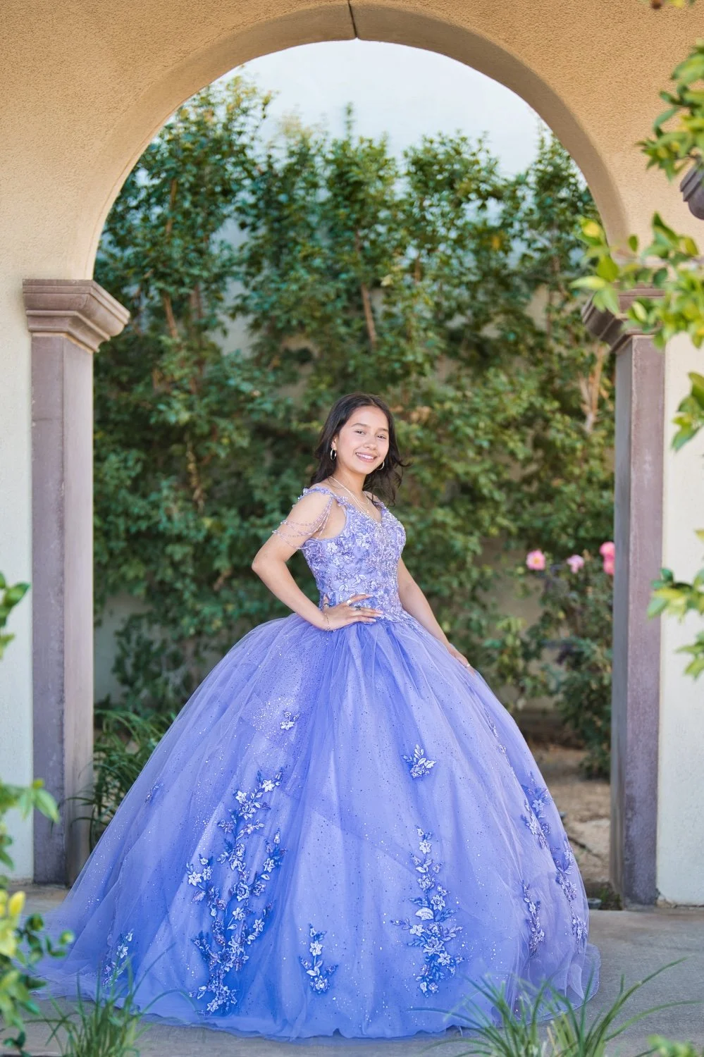 A girl in a purple quinceañera dress standing under an archway in a garden.
