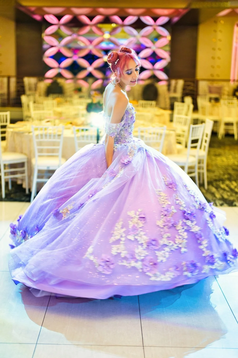A woman in a lavender ball gown with floral embellishments at a reception hall decorated with pink and purple lighting.