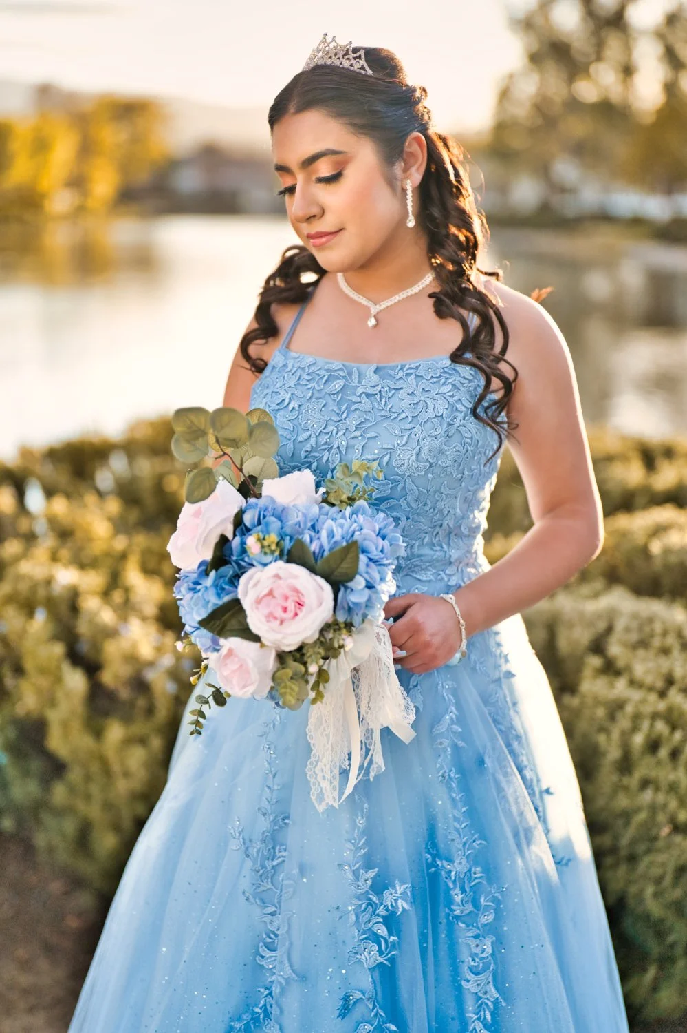 A young woman in a light blue lace dress holding a bouquet of pink and blue flowers, wearing a tiara and jewelry, outdoors near a body of water at sunset.