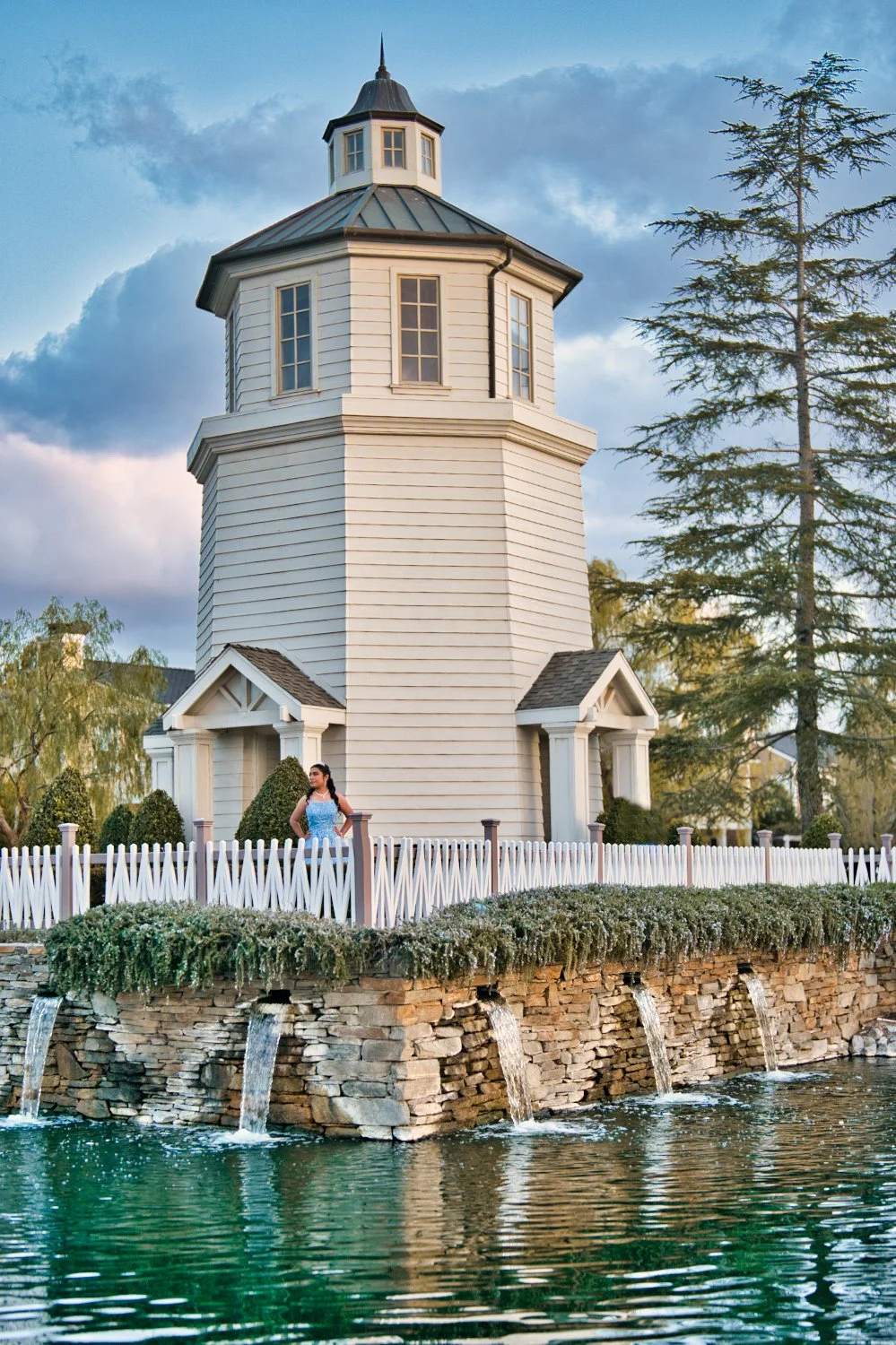 A white wooden lighthouse with a woman in a blue dress standing near a white picket fence next to a stone wall with small waterfalls and a body of water in the foreground. Tall trees and a cloudy sky are in the background.