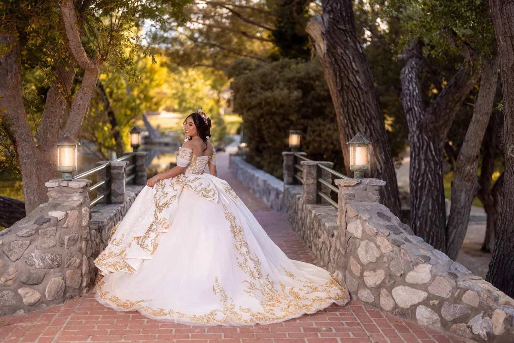 A woman in a white and gold ball gown standing on a stone and brick bridge, surrounded by trees with green foliage, during sunset.