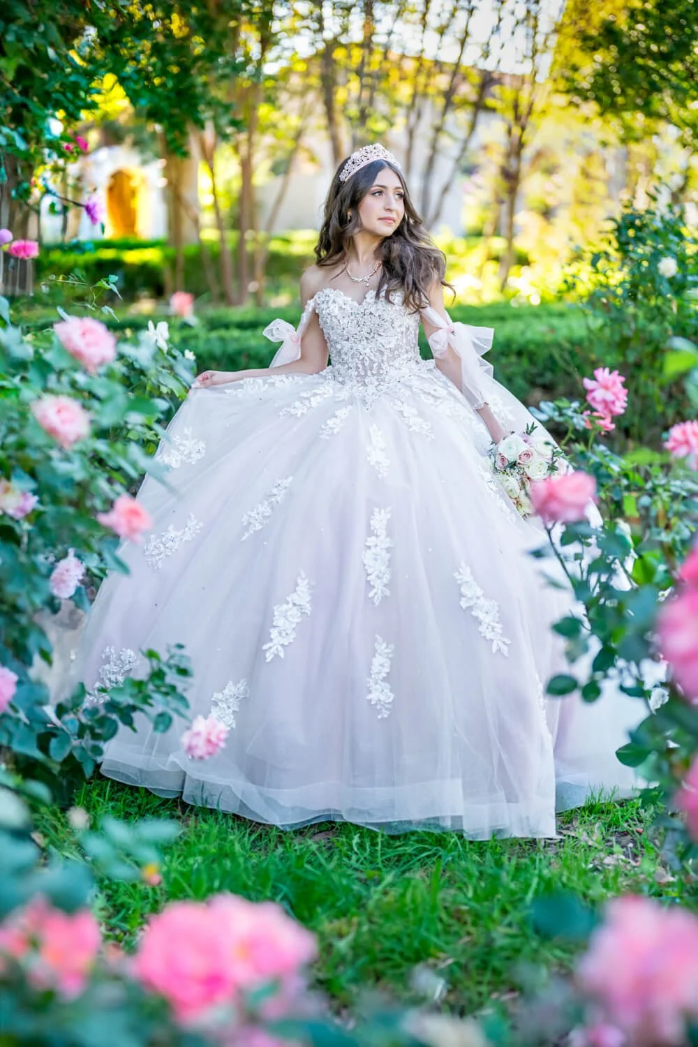 Bride in a white wedding dress with floral lace details, wearing a tiara, holding a bouquet, outdoors in a garden with pink roses and green trees.