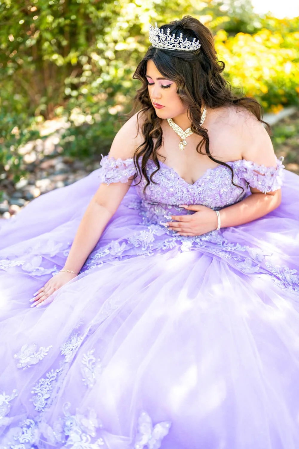 A woman in a lavender ball gown with lace and floral embroidery, wearing a tiara and jewelry, sitting outdoors in a garden.