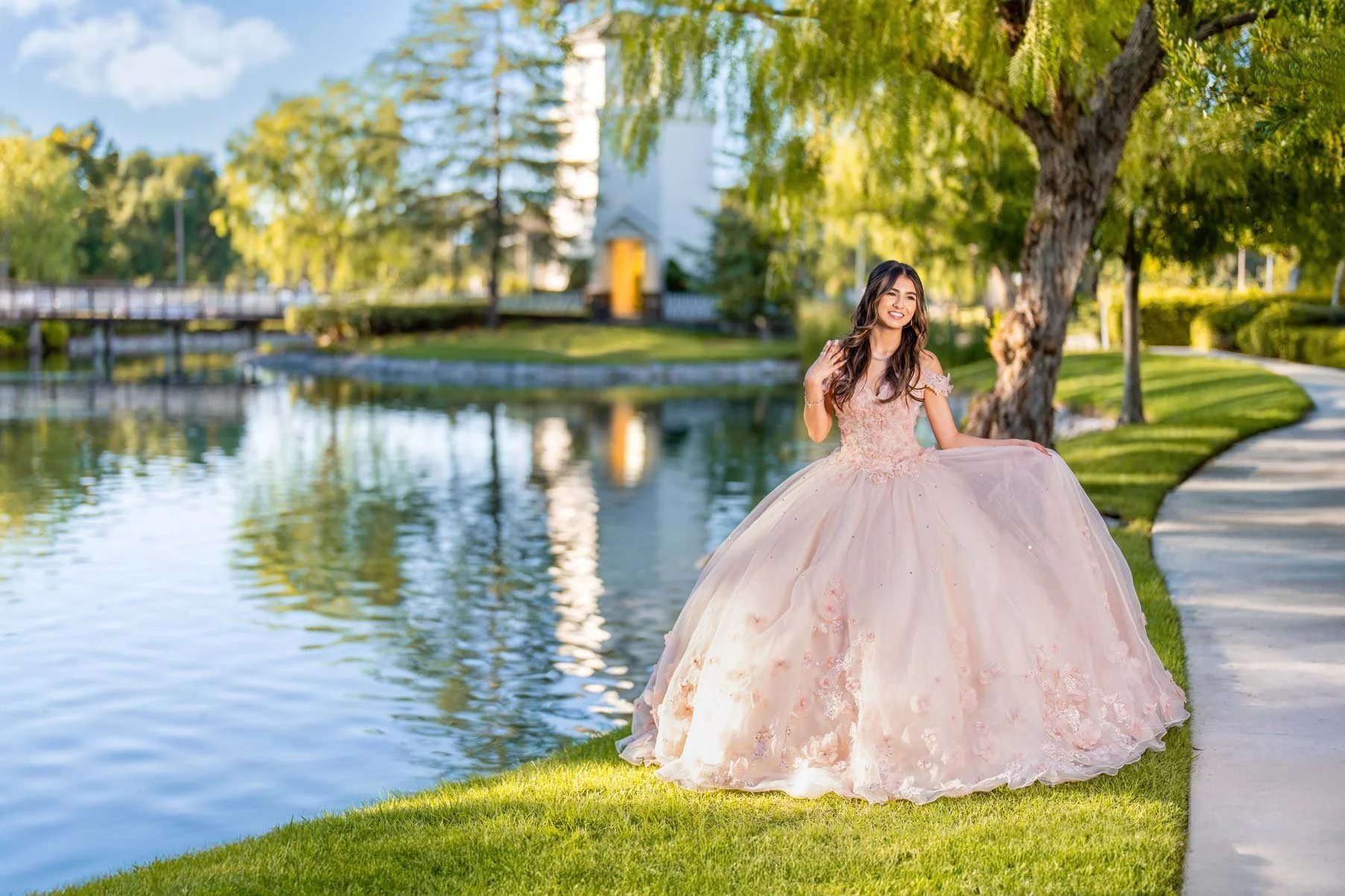 A young woman in a pink ball gown standing on a grassy edge by a pond in a park with trees and a house in the background.